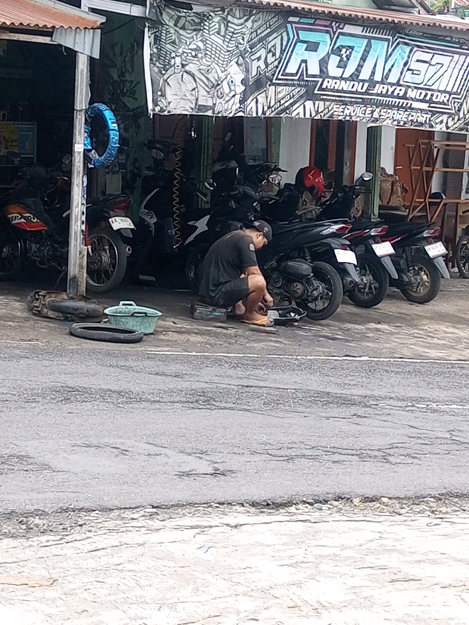 The image captures a scene outside a motorcycle repair shop in Keseneng, Indonesia. A man, dressed in a black t-shirt and shorts, is crouched on the ground, seemingly working on a motorcycle. He is wearing flip-flops. Several motorcycles are parked in the background, some of which are partially obscured by the man and other objects. A blue-wrapped tire is hanging from a pole on the left side of the image. A green plastic basin and a spare tire are on the ground in front of the man. A banner with text that appears to be in Indonesian hangs above the shop entrance. The shop is open-air, with motorcycles visible inside. The ground is paved with asphalt and appears slightly wet in places.