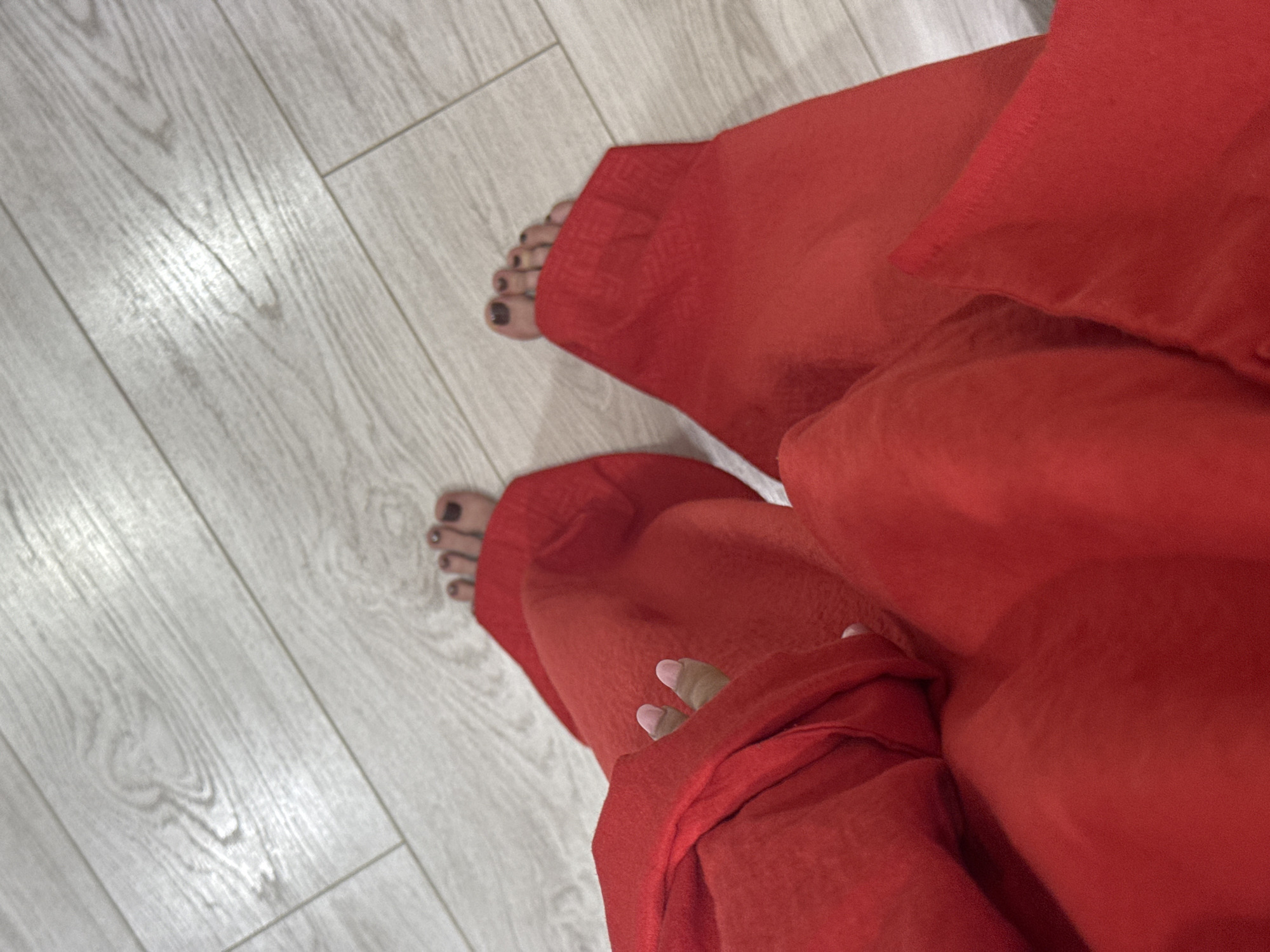 The image shows a view of someone standing on a light-colored, wood-patterned floor. They are wearing a voluminous, red garment that drapes around them, obscuring much of the lower body. The bare feet of the person are visible at the edges of the fabric, with dark nail polish on the toenails. The floor appears to be indoors and well-lit. No other specific details, such as the location in Izhevsk, Russia, can be discerned.