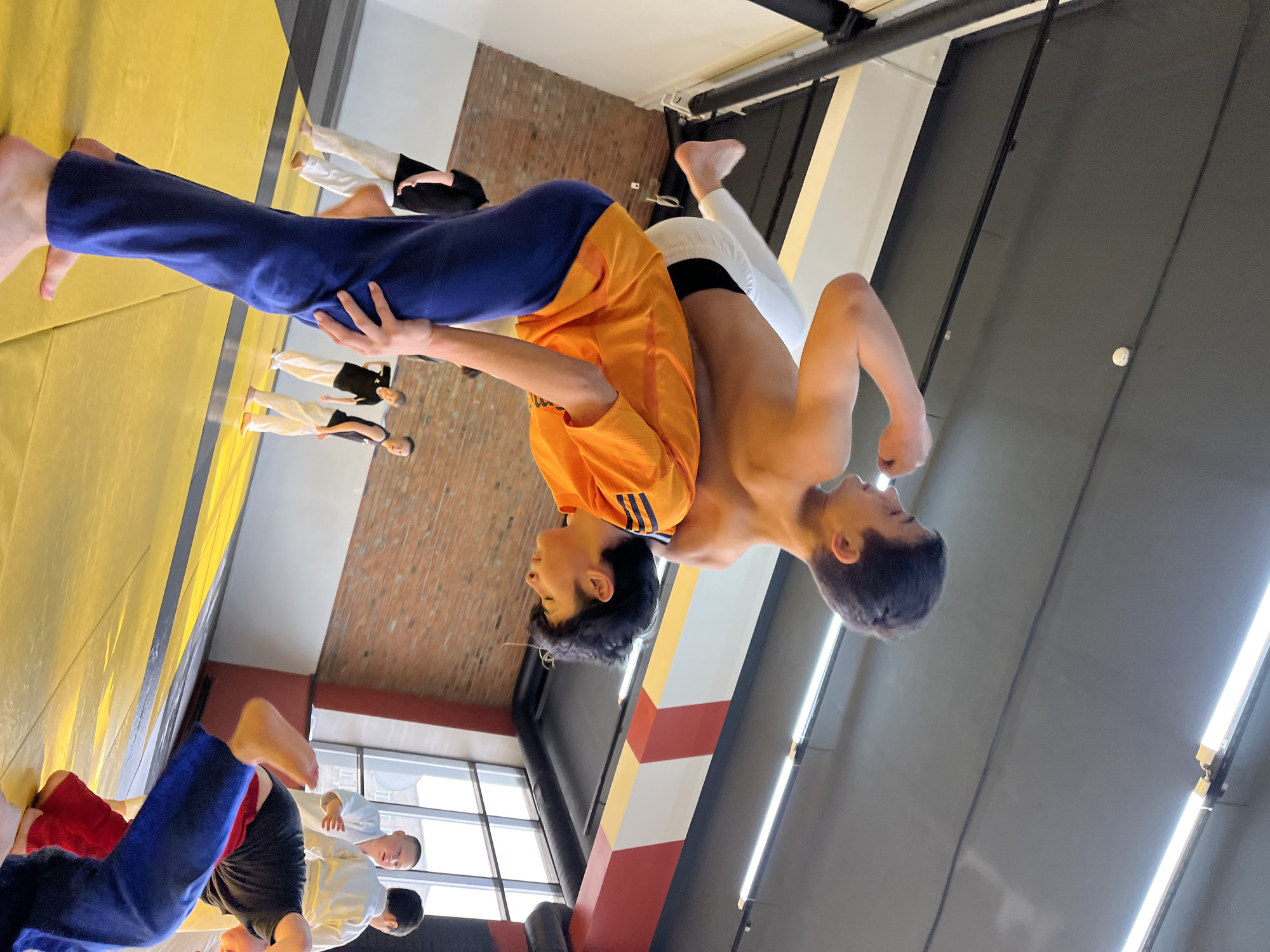 The image shows two young men engaged in a martial arts maneuver, possibly judo or a similar grappling sport. One man, wearing blue pants and an orange shirt with three stripes on the sleeves, is lifting the other. The second man is shirtless and wearing white pants with a black stripe at the waist. The surrounding environment includes a padded mat, likely in a gymnasium or training facility. Other people are visible in the background, some wearing similar martial arts attire. The location is identified as Krasnoyarsk, Russia, suggested by the picture's orientation and layout of the room. The image gives the feeling of action, balance, and athletic training.