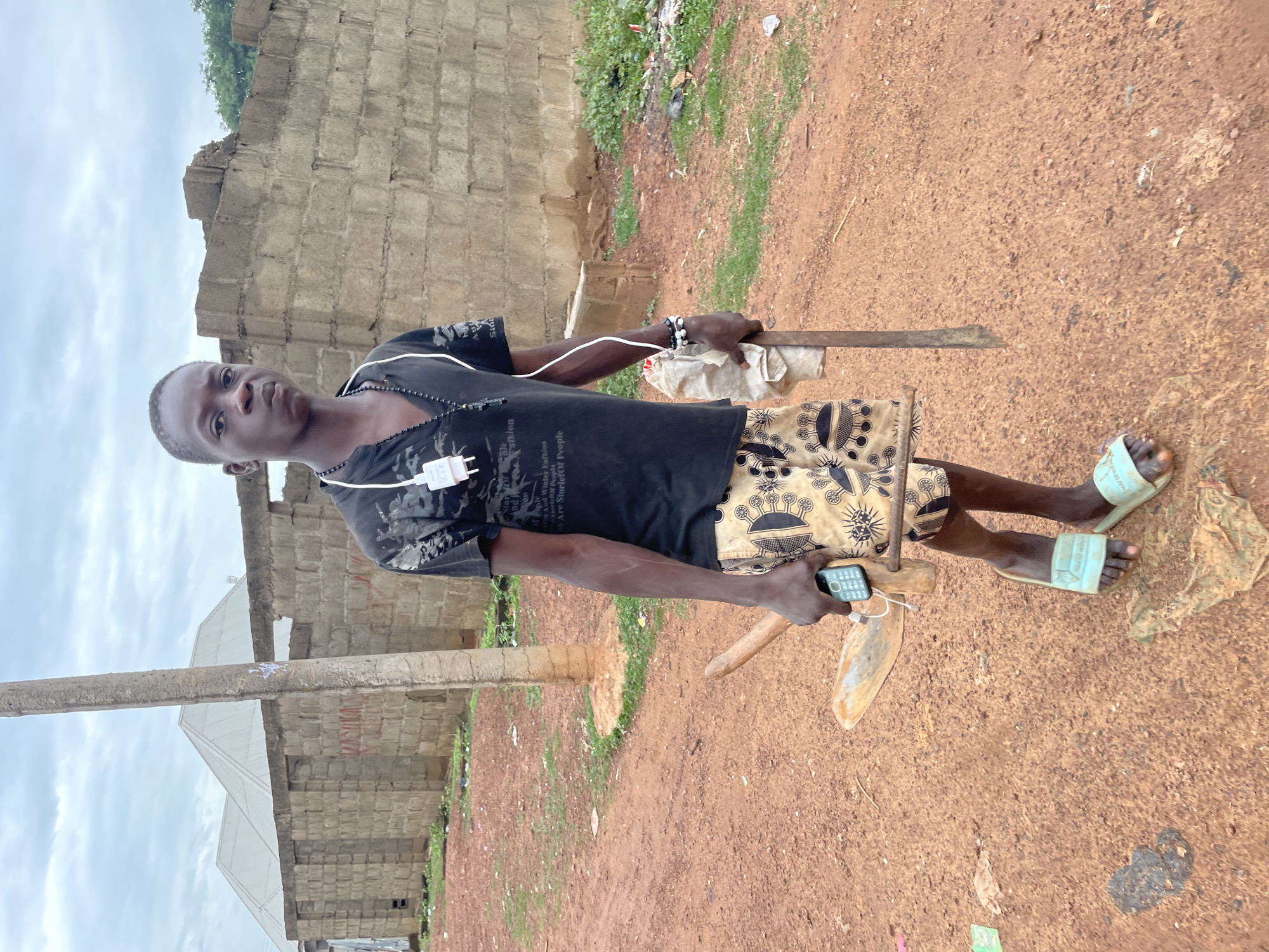 A young Black male stands outside in an overcast environment, holding a hoe and a machete, and a phone in his hand. He is wearing a black t-shirt, patterned shorts, and light blue flip-flops. The background features unfinished buildings made of concrete blocks. The ground is covered in dirt and some debris. The sky is cloudy.
