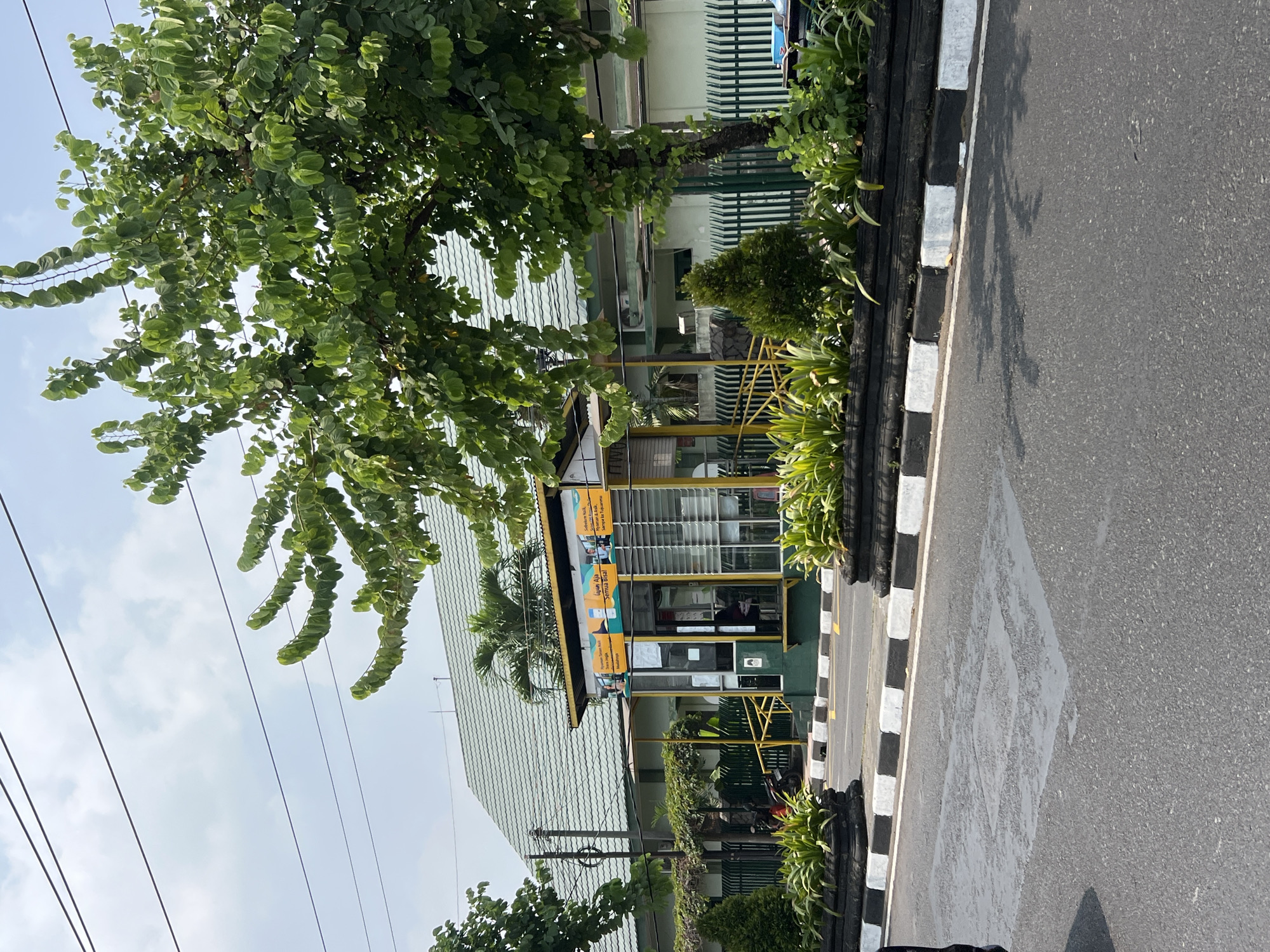 In the city of Yogyakarta, Indonesia, a bus stop is visible with a yellow roof and green pillars. The bus stop has glass walls and doors, and there is a person sitting inside. In front of the bus stop, there is a black and white striped curb and a road. Behind the bus stop, there is a building with a green roof and a fence. There are also trees and plants in the scene. The sky is cloudy.