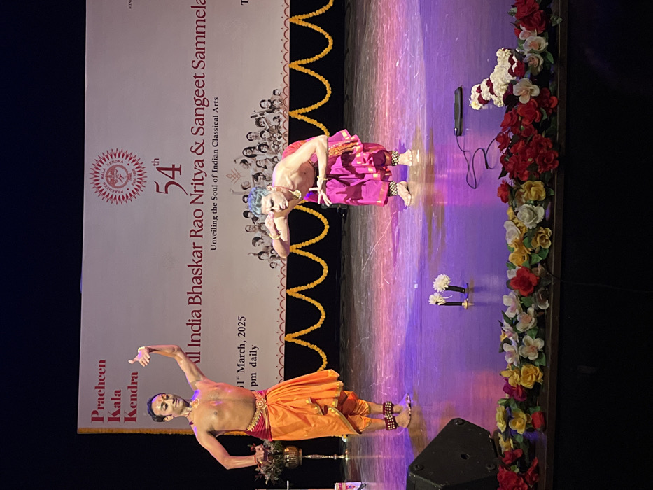 The image depicts a stage performance of Indian classical dance. Two dancers, one male and one female, are performing on a stage. The stage is decorated with garlands of colorful flowers. The male dancer is wearing a bright pink and yellow costume, while the female dancer is wearing a bright orange and red costume. Both dancers are barefoot. The male dancer is kneeling and extending his left hand towards the audience. A banner behind the dancers reads "Pracheen Kala Kendra" and "54th All India Bhaskar Rao Nritya & Sangeet Sammela Unveiling the Soul of Indian Classical Arts". The performance appears to be taking place at Pracheen Kala Kendra, a cultural center located at the given coordinates (latitude: 30.733, longitude: 76.789), in Sector 35, Chandigarh, India. The floor of the stage is polished wood. A microphone stand and a few flowers can be seen at the front of the stage. The stage is illuminated by spotlights. The background is dark. The image appears to be a still from a video recording of the performance.