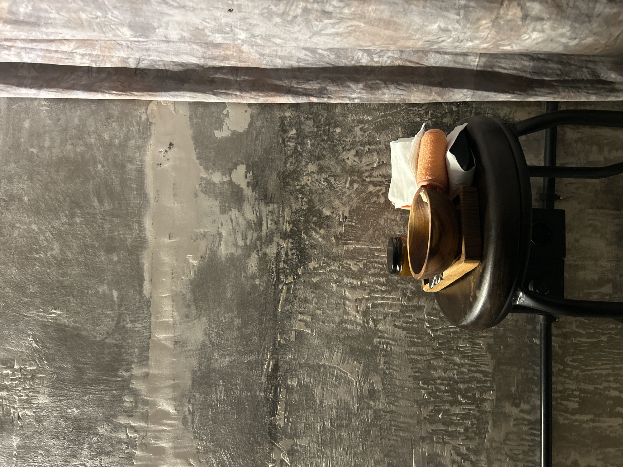 A close-up, eye-level shot captures a dark, textured wall in North Kuta, Indonesia. To the right, a patterned curtain hangs vertically, its folds creating shadows. In the center, a dark metal stool supports a wooden board. On the board rests a wooden bowl, a small glass jar with a black lid, a folded orange towel, and white folded napkins. The wall's surface is rough and uneven, with patches of lighter and darker gray, suggesting a distressed or concrete finish. The lighting is dim, casting subtle shadows on the stool and its contents. No people are visible in the scene.