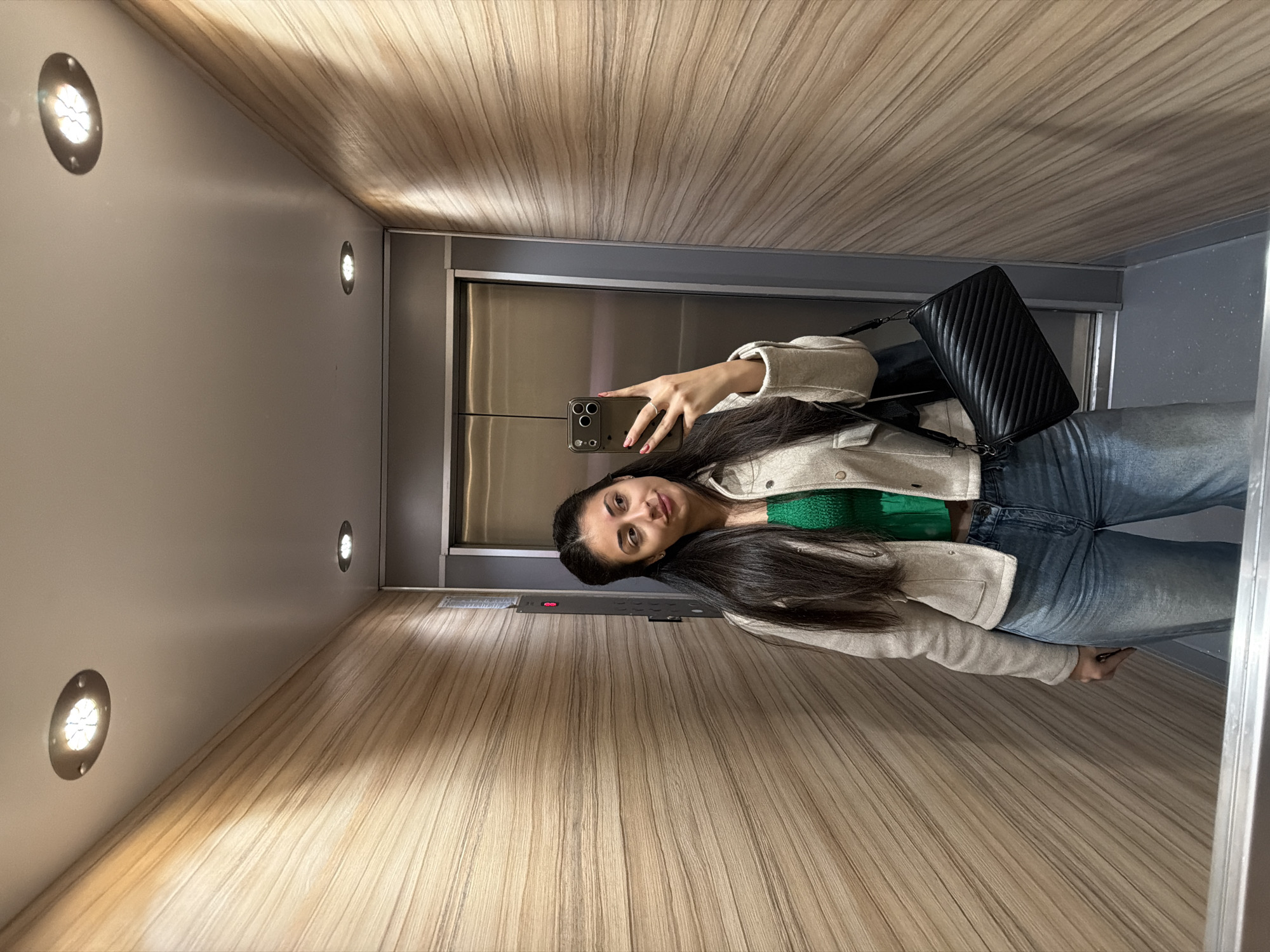 A young woman takes a mirror selfie inside an elevator. The elevator's interior has wooden-paneled walls and a grey metal frame for the door. The woman is wearing blue jeans, a green top, and a light-colored jacket. She holds a phone in her right hand to capture the image. A black purse hangs over her left shoulder. Several round recessed lights are visible on the elevator's walls. The location is implied to be in Sofia, Bulgaria, but there is no direct evidence in the image to confirm the exact location.