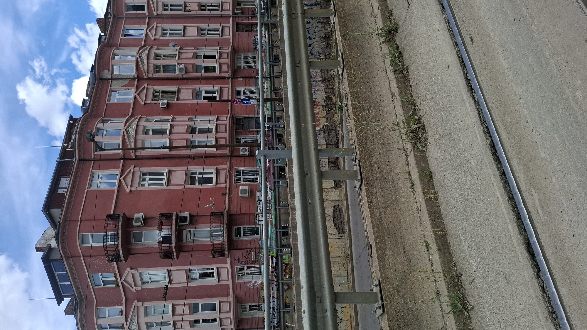 This is a wide shot photograph taken on a cloudy day, capturing a section of a city street in Sofia, Bulgaria. The dominant subject is a multi-story pink building with an ornate facade featuring bay windows and balconies. The building appears to be of older architecture, with visible air conditioning units and satellite dishes on its exterior. Below the building, a metal guardrail runs horizontally across the frame, separating the street from a concrete embankment. This embankment is cracked and overgrown with sparse vegetation. In the foreground, a tram track is visible embedded in the concrete road surface. The overall impression is one of an urban environment, possibly a slightly neglected area of the city, with signs of everyday city life such as the guardrail and tram tracks.