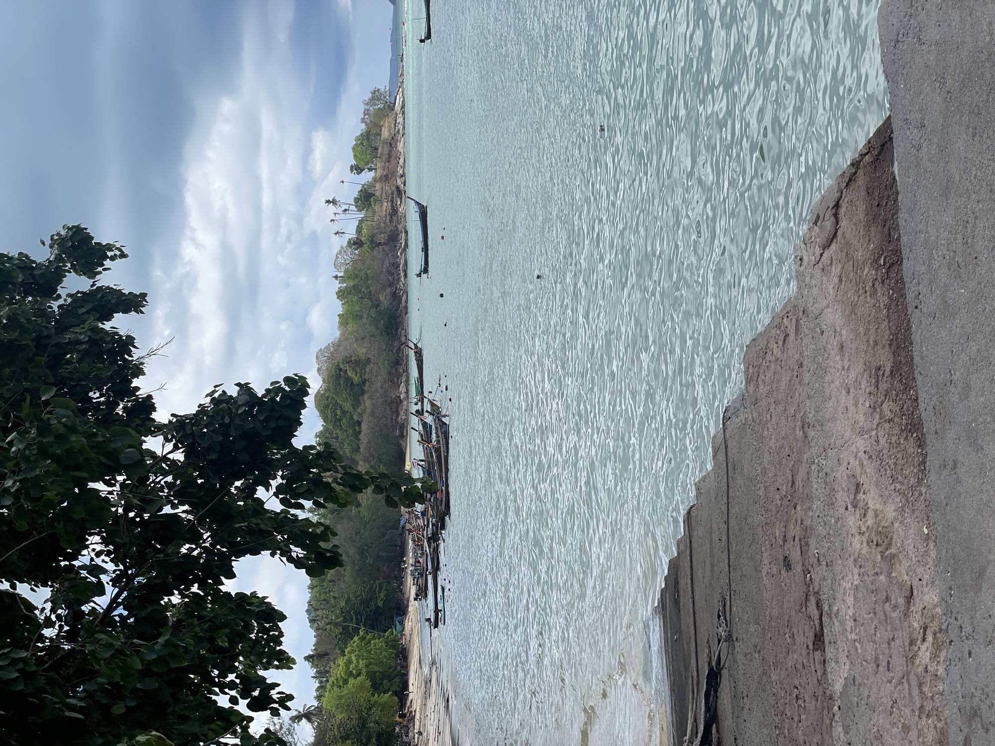 The image captures a coastal scene in Rawai, Thailand, featuring a concrete structure in the foreground from which the photograph appears to be taken. The clear, light-blue water extends towards the shoreline, where numerous traditional Thai longtail boats are docked. Lush green vegetation covers the land adjacent to the beach, interspersed with palm trees. The sky is overcast with cloudy conditions, casting a muted light over the scene. No people are visible in the frame. A tree with dense foliage is located to the left of the frame.
