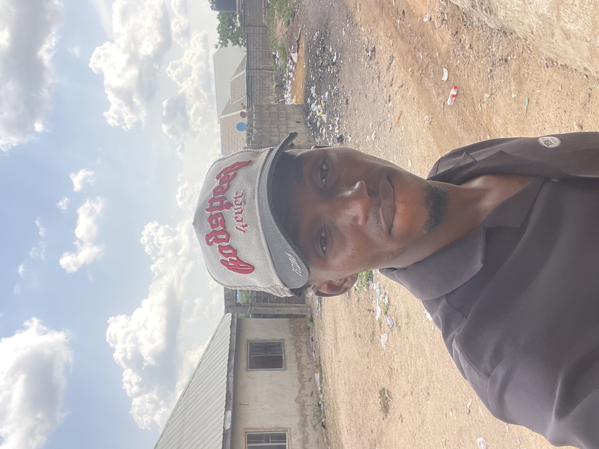 A selfie shows a man wearing a cap with the word "Speed" printed on it in red. The man is located in an outdoor environment in Lokoja, Nigeria. He is standing on an unpaved road or dirt path. Debris and litter are scattered across the ground and in the background. Partially constructed buildings are visible, with gray brick. The sky is light blue with a scattering of fluffy white clouds.