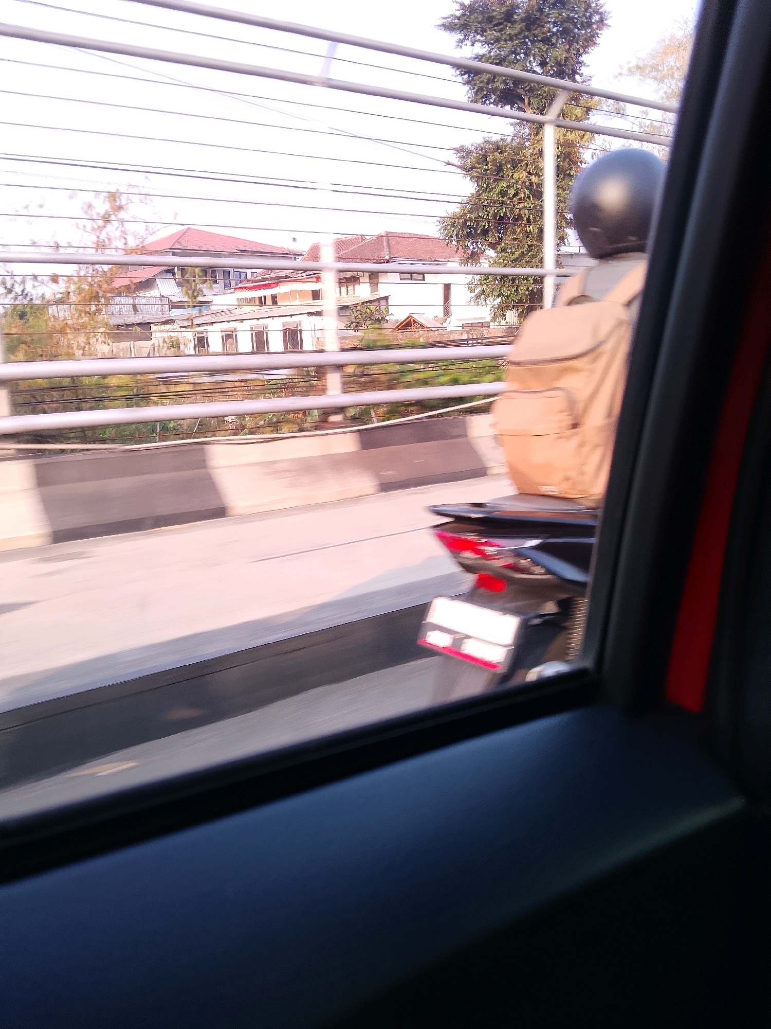 The image, captured from the interior of a vehicle, shows a view through a window looking out onto a street scene. In the foreground, a dark interior with a red accent is visible, suggesting the passenger side of a car. The primary focus of the exterior view is a motorcycle rider wearing a black helmet and a tan backpack, seen from behind. The motorcycle has a black body and red taillights. It is traveling on an elevated roadway, indicated by the concrete barrier and railing.
Beyond the roadway, several buildings with red-tiled roofs are visible, characteristic of residential architecture. The sky is bright, suggesting daytime, possibly late afternoon due to the warm lighting. A large tree with green foliage stands to the right of the frame, behind the motorcycle rider. The overall impression is a snapshot of daily life in a moderately urban environment. The specific location in Malang, Indonesia, is not explicitly identifiable from visual cues within the image, though the architectural style and vegetation are consistent with tropical Southeast Asian settings. The motion blur suggests the vehicle from which the photo was taken was in transit.