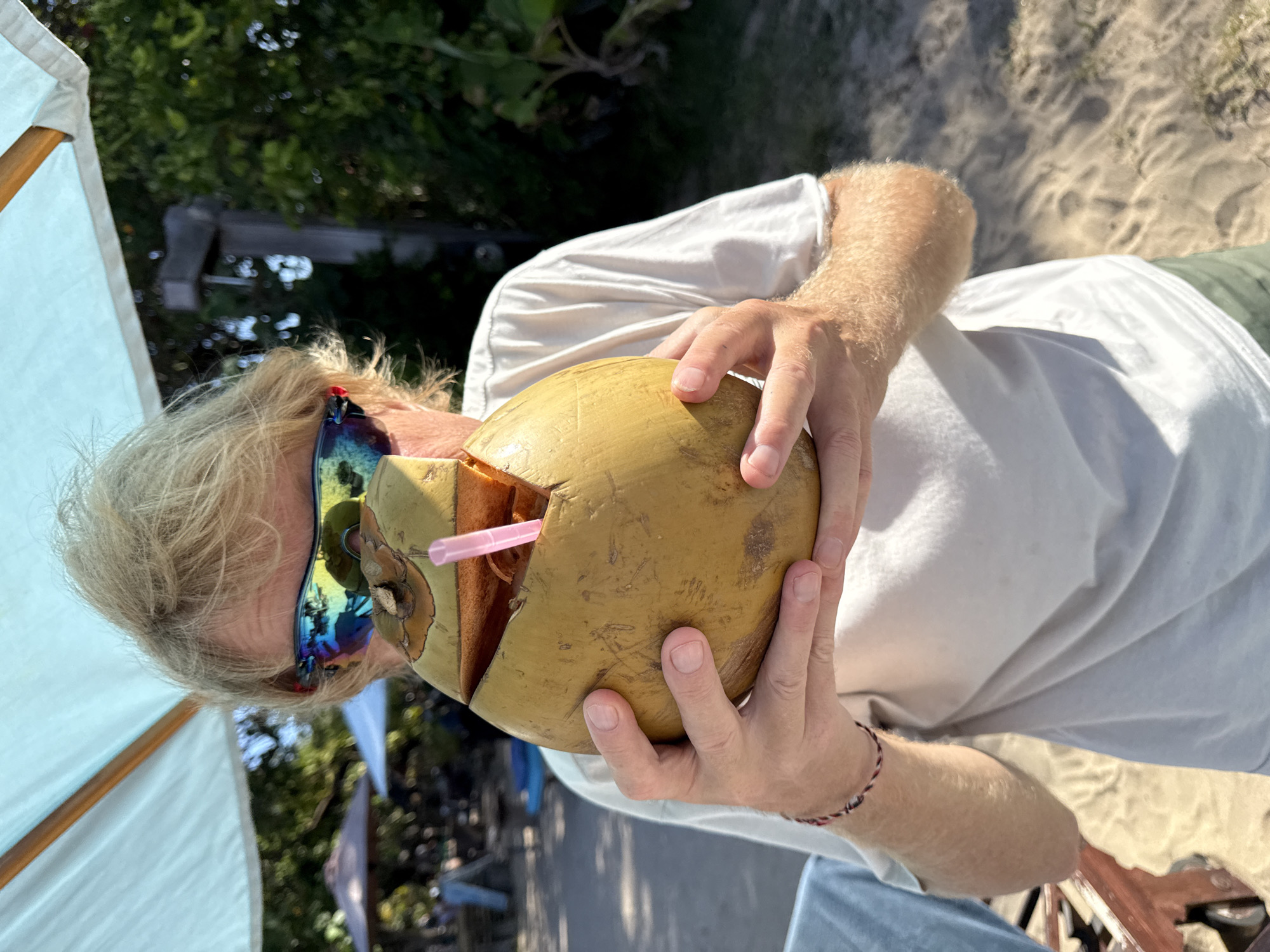 A person with blonde hair and sunglasses is holding a large, halved coconut with a pink straw in it. They are wearing a white t-shirt and green shorts. The person is standing on a sandy beach, with trees and umbrellas in the background. The scene appears to be outdoors in a tropical location, possibly Denpasar, Indonesia, given the presence of lush vegetation and beach umbrellas. The lighting suggests it is daytime. The person is looking down at the coconut. The image is taken from a medium close-up perspective.