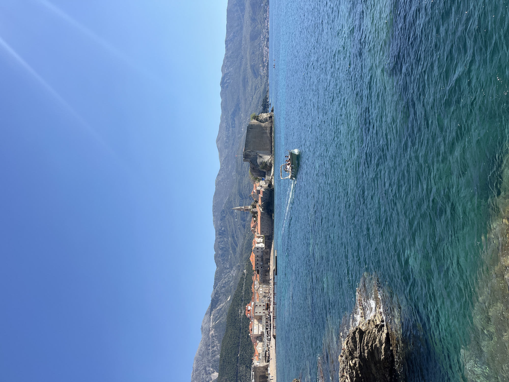 The image shows the city of Budva, Montenegro from a viewpoint near the sea. In the background, there are large mountains with lush green vegetation. In the center, there is a medieval town characterized by terracotta rooftops, white stone buildings, and a prominent church spire. A stone fortress is situated on a promontory extending into the sea. In the foreground, the clear turquoise water of the Adriatic Sea fills the right side of the image. A small boat with passengers is visible on the water near the medieval town. Rocky terrain is present at the bottom right corner. The sky is clear blue with a bright light source at the top left.