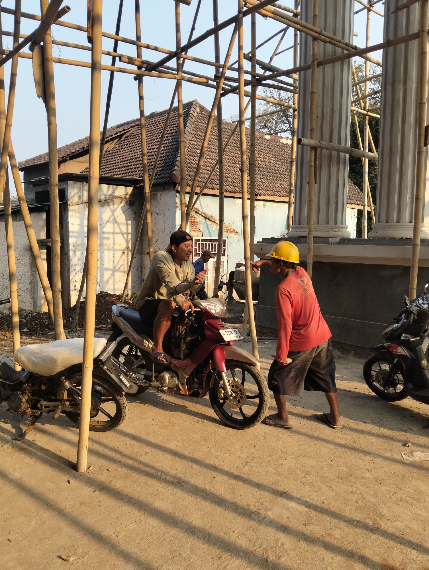 In Adinuso, Indonesia, on a construction site with bamboo scaffolding and partial columns, two men are engaged in conversation. One man, wearing a light-colored shirt and a dark baseball cap, is seated on a maroon and silver motorcycle. He is gesturing with his right hand, holding a dark object, possibly a phone. The second man, dressed in a red long-sleeved shirt and dark shorts, stands facing the seated man. He wears a yellow hard hat and is pointing with his left hand. Two motorcycles are visible in the scene, one maroon and silver, and another black one to the right. The ground is unpaved and dusty. In the background, a house with a tiled roof and a partially constructed building with columns are present. The lighting suggests it is either early morning or late afternoon.