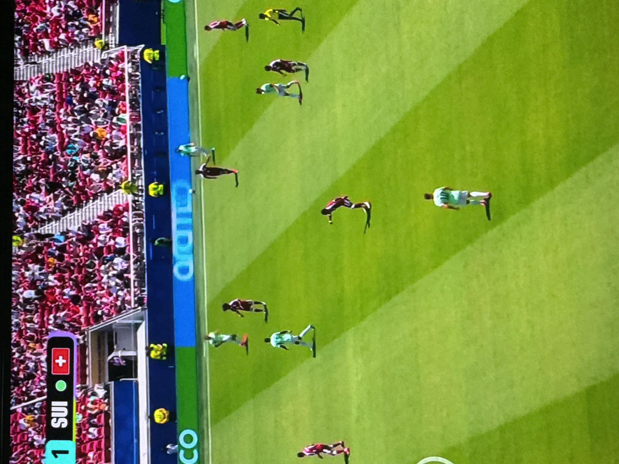 An aerial view captures a soccer match on a sunlit, bright green field with distinct parallel mowing patterns. In the upper left corner, a scoreboard displays "1 SUI" alongside a green circle and a Swiss flag icon, indicating the score and potentially the team. Spectators dressed in red and white fill the stadium seats in the background. Several players are visible on the field; one player in a light green jersey with the number 10 on the back is walking away from the camera towards the right side of the frame. Another player in a maroon jersey with white trim is facing the camera and appears to be kicking a ball. A player in a light green jersey is running on the field, and another in a maroon jersey is standing near him. Further down the field, a player in a maroon jersey is running. Several other players in maroon and light green jerseys are scattered across the field. The environment is an outdoor sports stadium.
