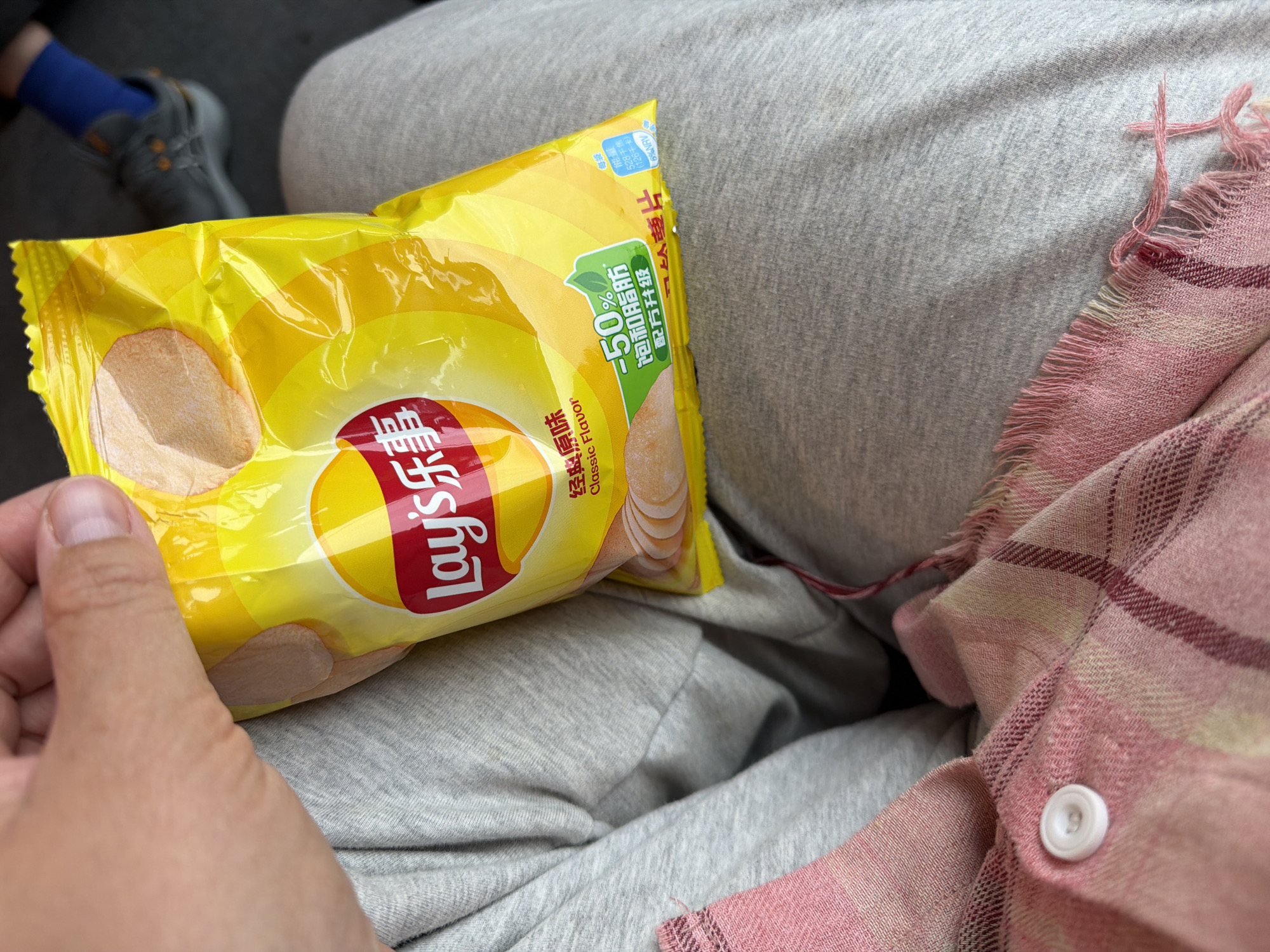 The image shows a close-up of a person holding a bag of Lay's Classic Flavor potato chips. The person is wearing gray sweatpants and a pink plaid scarf with a white button. The bag of chips is yellow with red lettering and depicts images of potato chips. Some text in Chinese characters is visible on the bag, as well as a green graphic indicating "-50% less fat and oil". In the background, a blue sock and a gray sneaker are visible. The scene appears to be indoors, possibly in a vehicle or at a public transport hub, given the casual clothing and the glimpse of footwear. The location in the city of Khimki, Russia, is not directly indicated by any visual cues in the image.