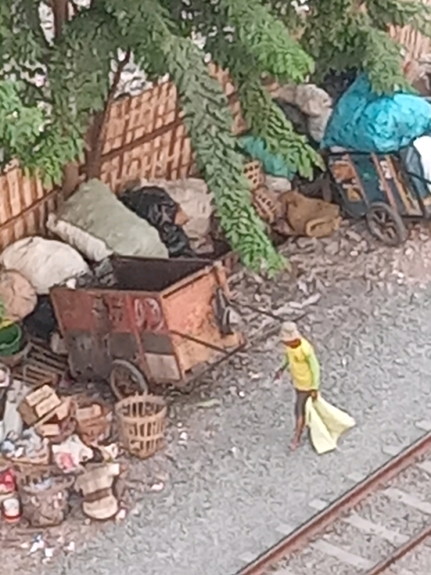 The image depicts a person walking along a railway track in an urban environment, likely a disposal or scrap area, near Surabaya, Indonesia. The ground is covered in gravel and debris, with various piles of waste and discarded items. In the foreground, a set of railway tracks runs diagonally across the lower right portion of the frame. A person, wearing a yellow shirt and a light-colored hat, is walking away from the camera, carrying a yellow bag or cloth in their right hand. To the left of the person, there is a rusty, orange cart with wheels, filled with debris. Behind the cart, there are several large piles of materials, including what appear to be sacks of fabric, some plastic sheeting, and other unidentifiable objects. A wooden fence, partially covered by green foliage, runs along the upper left section of the image. The overall scene suggests a working or scavenging area within the city.