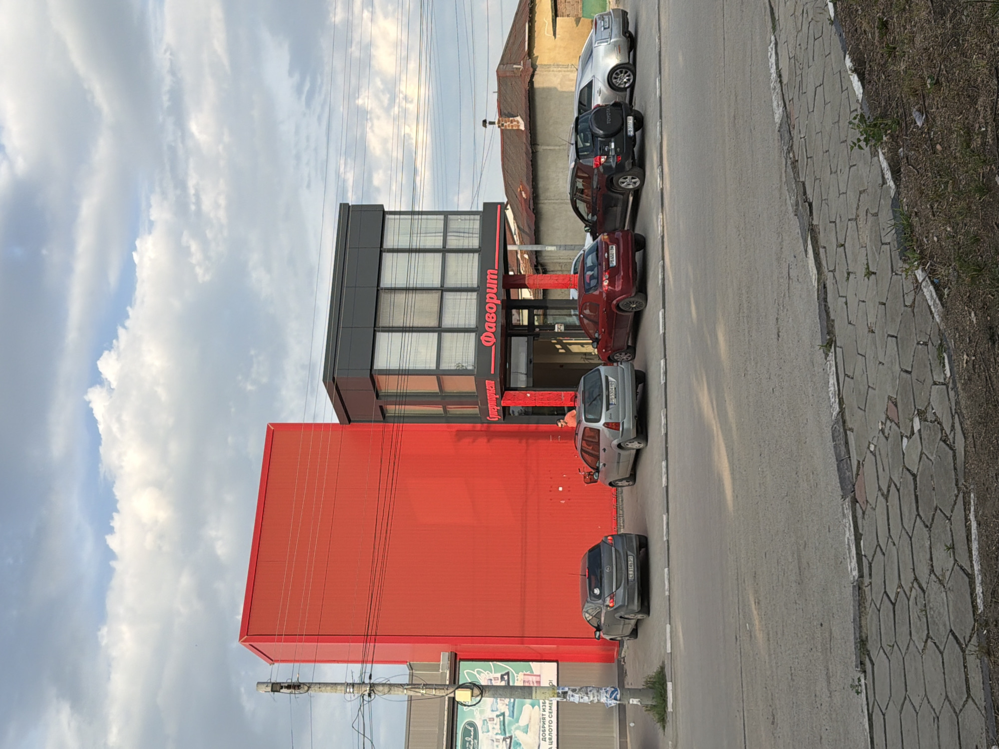 This image captures a street scene in Saedinenie, Bulgaria, featuring a modern commercial development adjacent to an older residential structure. In the foreground, a gray asphalt road runs horizontally, bordered by a curb and a sidewalk paved with hexagonal gray concrete blocks, interspersed with sparse weeds.
The central focus is a multi-part commercial building. To the left, a tall, plain, rectangular structure is clad entirely in bright red corrugated material. Immediately to its right is a two-story building with a dark gray facade and prominent windows on the upper floor. This building features a red overhang with the Cyrillic text "Фаворит" (Favorit) in white script, indicating a store or supermarket. Below "Фаворит," another sign reads "Супермаркет" (Supermarket). A red support column stands at the entrance.
Parked in front of the "Фаворит" building are several cars. From left to right, visible are: a dark gray Opel Astra hatchback with license plate "CT 1146 PT," a silver station wagon with license plate "PB 5304 HB," a red Chevrolet Lanos/Aveo sedan with license plate "PB 6001 BA," a black Toyota RAV4 SUV with a rear-mounted spare tire and license plate "PB 3117 TE," and a silver Cadillac sedan.
To the far left, partially visible, is another building with a green billboard featuring white text in Bulgarian, including "ДОБРИЯТ ИЗБОР ЗА ЦЯЛОТО СЕМЕЙСТВО" (The good choice for the whole family) and imagery of packaged goods. A gray utility pole with various attached wires stands prominently in front of this billboard. Overhead, numerous power lines stretch across the scene.
A single person, a male in an orange t-shirt, is visible standing near the entrance of the "Супермаркет."
Behind the commercial buildings, an older, single-story house with a weathered tiled roof and a brick chimney is visible. The sky above is mostly overcast with scattered white and gray cumulus clouds, suggesting either late morning or early afternoon with soft, diffused sunlight. The overall impression is a mix of contemporary commercial development and older infrastructure in a suburban or semi-urban setting.