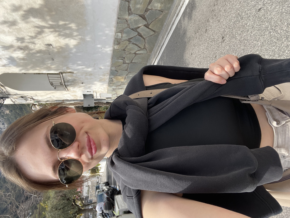 A female subject is depicted in a close-up outdoor portrait, standing on a paved street in what appears to be the city of Arienzo, Italy, under clear daylight conditions. She has light skin, brown hair, and wears round, gold-framed Ray-Ban sunglasses with dark lenses, reflecting the environment. Her lips are tinted light pink, and she exhibits a faint smile. Her attire consists of a black sleeveless crop top, light beige corduroy trousers, and a dark grey sweatshirt tied around her shoulders and neck. A light-colored shoulder bag strap is visible over her right shoulder, and her right hand grips the cuff of the sweatshirt. The background shows a descending paved street with parked vehicles, including a scooter and cars, on the left. On the right, a building features a light-colored upper facade with foliage shadows and a stone-clad base. An arched alcove with a small wrought-iron balcony is present on an upper level, along with utility cables. Distant green hills or mountains are visible under a clear sky.