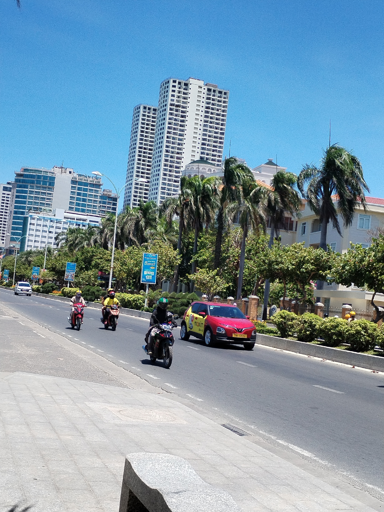 The image captures a street scene in Nha Trang Ward, Vietnam. The sky is clear and blue. A four-lane road runs across the foreground, with vehicles, including a red and yellow Maxim taxi with license plate 75H-109.14 and several motorcycles, in motion. Three riders are visible on the motorcycles, two wearing bright colors (red and yellow) and one clad in black.
In the background, there are several high-rise buildings and hotels. Palm trees line the street along with other trees and bushes separating the road from houses with red-tiled roofs. On the sidewalk to the left is a grey bench. Blue advertising posters are visible next to the road, and there's a white car further down the street.