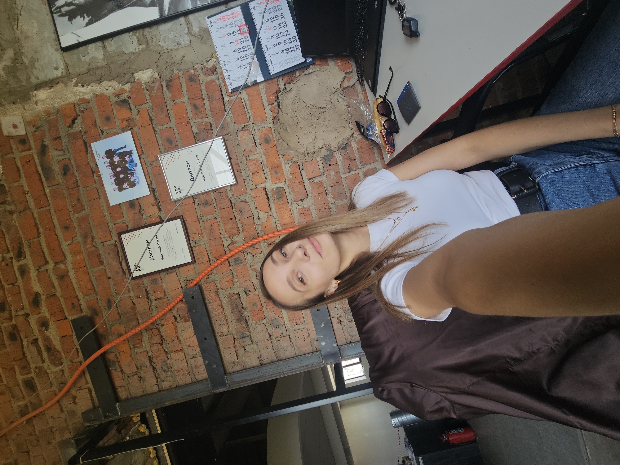 A woman is taking a selfie in a room with exposed brick walls. Several certificates and a group photograph are mounted on the wall. A calendar, laptop, sunglasses, and other small objects are on a desk. The image includes text indicating the location as Chaikovsky, Russia, and references to "Диплом" (Diploma) with the names "Фотиной Марии" (Fotinoy Marii) and "Тарасовой Елене" (Tarasovoy Elene).