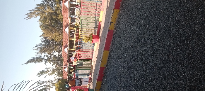 The image shows a view of a paved area in front of a row of buildings, likely a resort or hotel, in Athi River, Kenya. The buildings are yellow and white with red tile roofs. There is a colorful metal fence in front of them, with a small green bush and red flower pot. Three people are walking away from the camera towards the fence, and a fourth person is walking in the opposite direction. The ground is covered in small gravel, and there is a paved sidewalk with yellow and red markings. The trees in the background are green and lush. The sky is clear and blue.