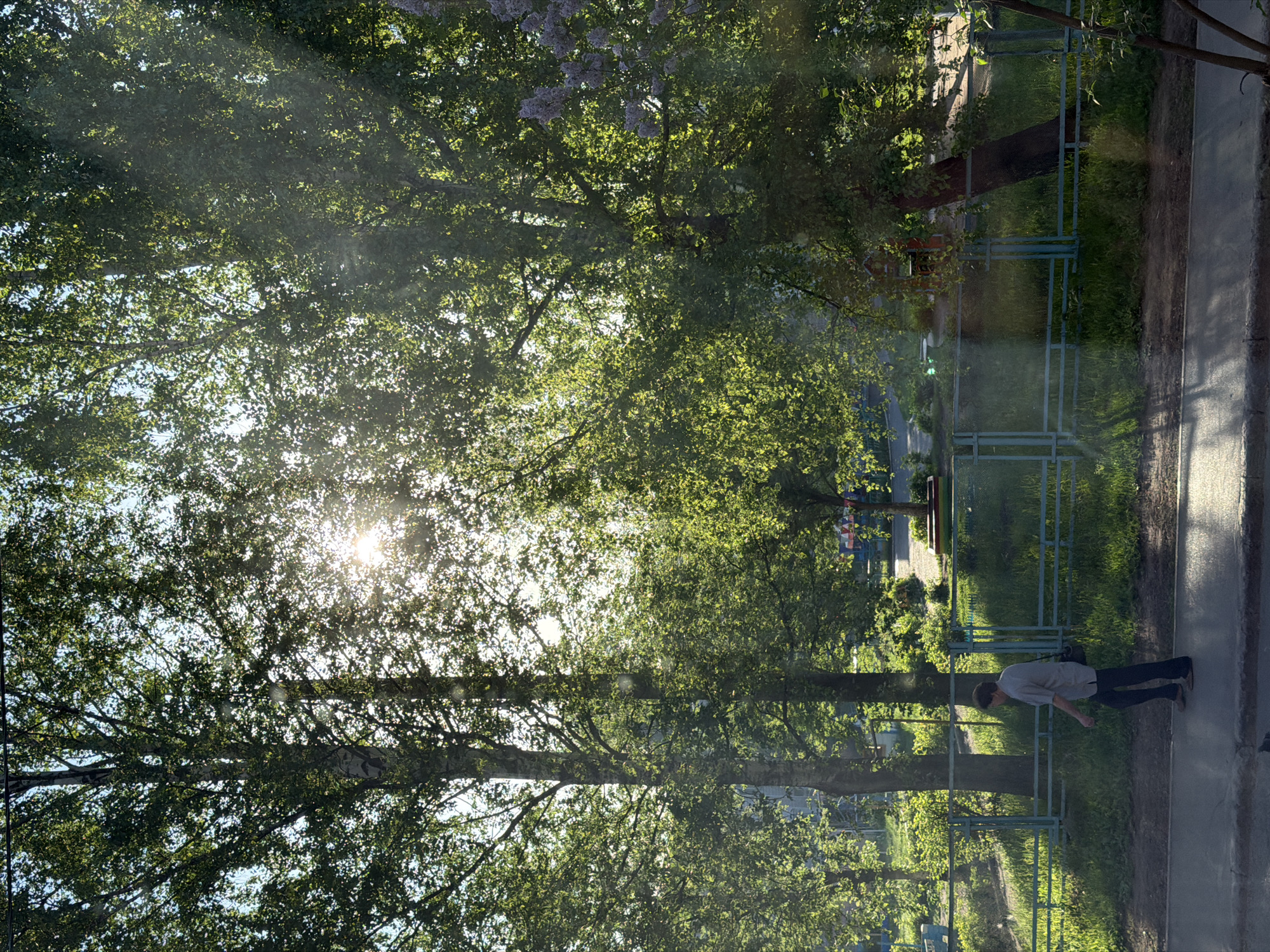 The image shows an outdoor scene in Izhevsk, Russia. Sunlight is visible through the foliage of trees on the left side of the frame. A sidewalk occupies the right portion of the image, where a person is walking. A fence separates the sidewalk from a grassy area with more trees and possibly a playground in the distance.