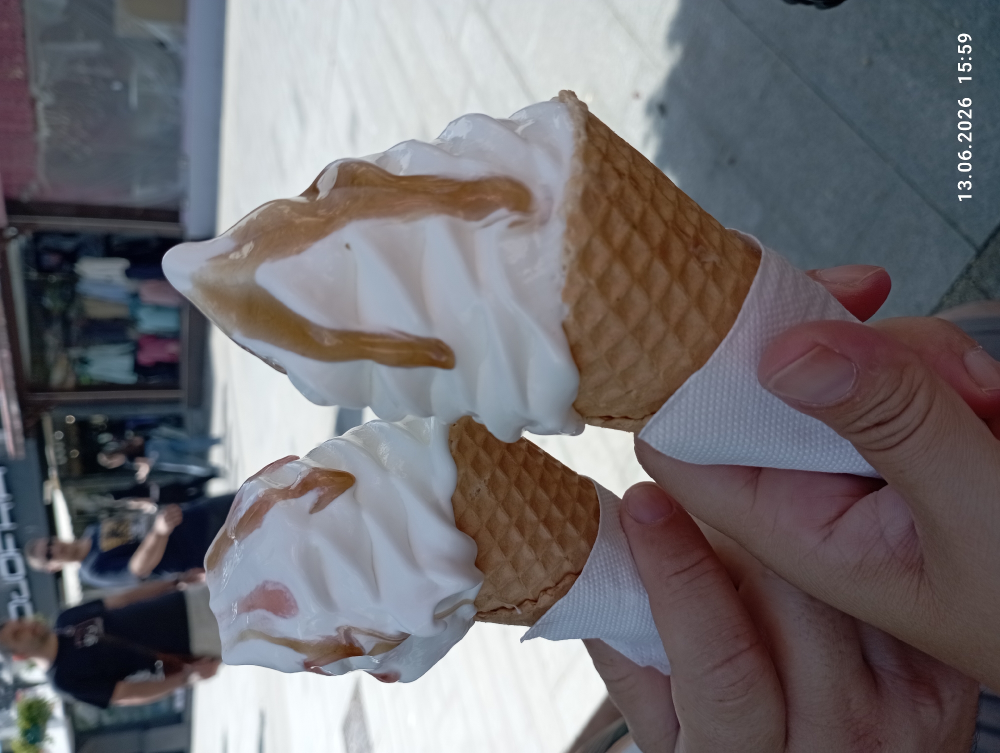 The image captures a close-up view of two ice cream cones held by two individuals, whose hands are visible. The ice cream appears to be soft-serve, with a white base and drizzled with a caramel or syrup topping. Both cones are wrapped in white paper. In the background, several people are blurred, suggesting an outdoor setting in a public area. The environment appears to be a street or walkway, with shops visible in the distance. The presence of the shops and the general atmosphere suggest an urban location. The timestamp at the bottom right corner indicates the image was taken on June 13, 2026, at 15:59. Based on the details, this scene is consistent with a street view in the city of Burgas, Bulgaria, possibly near a commercial area or promenade.