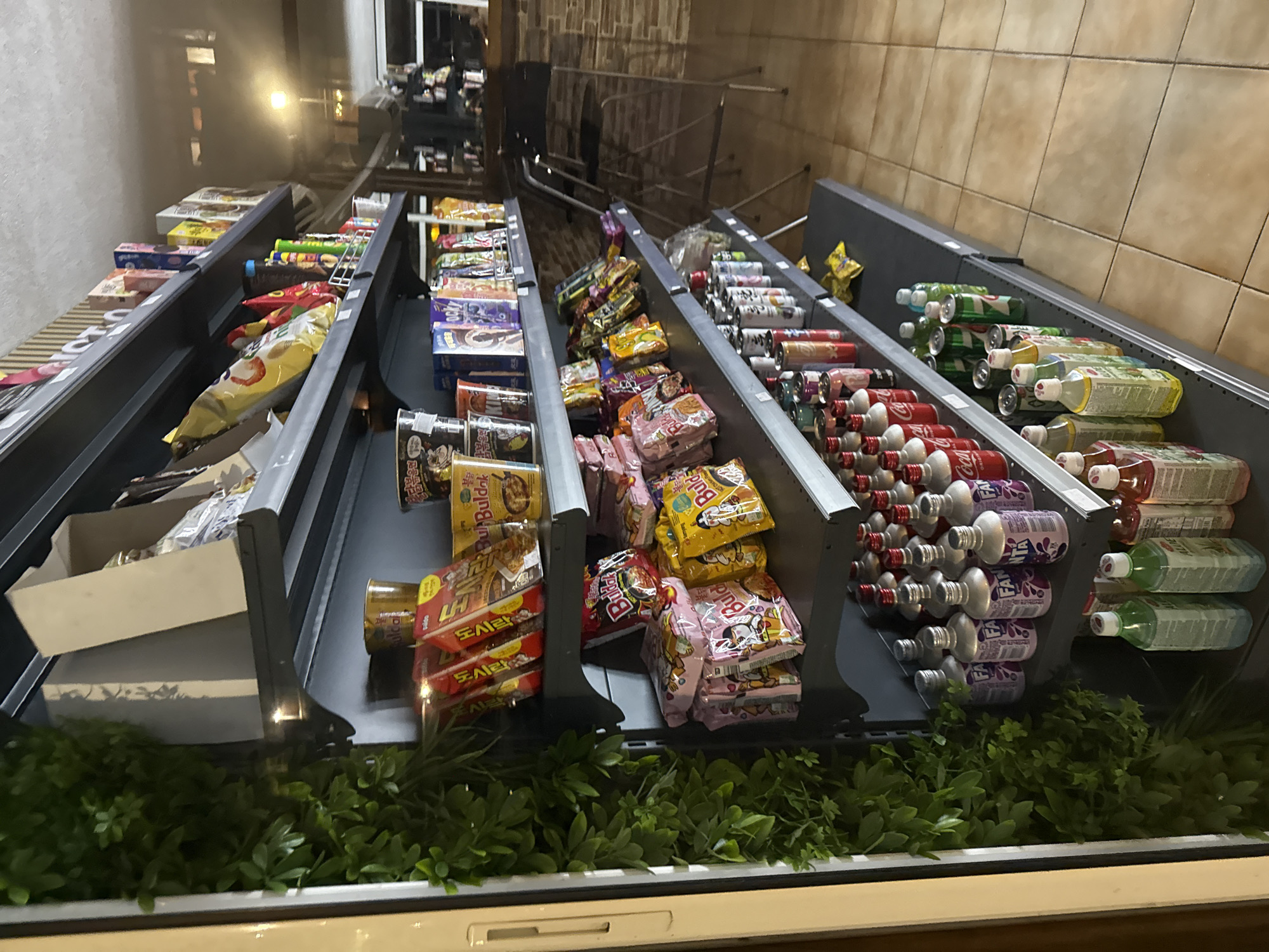 The image shows a series of tiered shelving units in what appears to be a convenience store or market in Varna, Bulgaria. The shelves are stocked with various items including chips, boxes of noodles, and bottles of beverages such as soda. The products are arranged in rows, filling the metal-framed shelves. Some artificial greenery is visible at the base of the shelves, adding a decorative touch. The floor has a light-colored tile pattern. The environment suggests an indoor commercial setting. In the background, there's some visible street and some architectural structures.