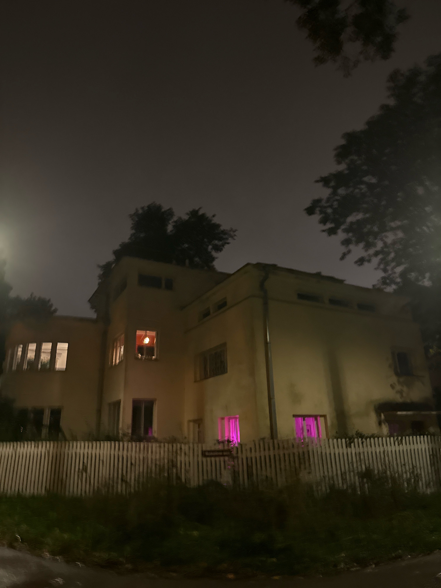 This image depicts a modern, two-story building at night, located in the Mustamäe district of Tallinn, Estonia. The building is illuminated by interior lights, some of which emit a pink or purple glow, suggesting decorative or ambient lighting. The exterior is a light-colored stucco. A white picket fence runs along the front of the property, with overgrown grass and vegetation visible between the fence and the street. Trees with dark foliage are silhouetted against the dimly lit, overcast sky. There are no people visible in the image. The overall impression is one of a quiet, residential nighttime scene.