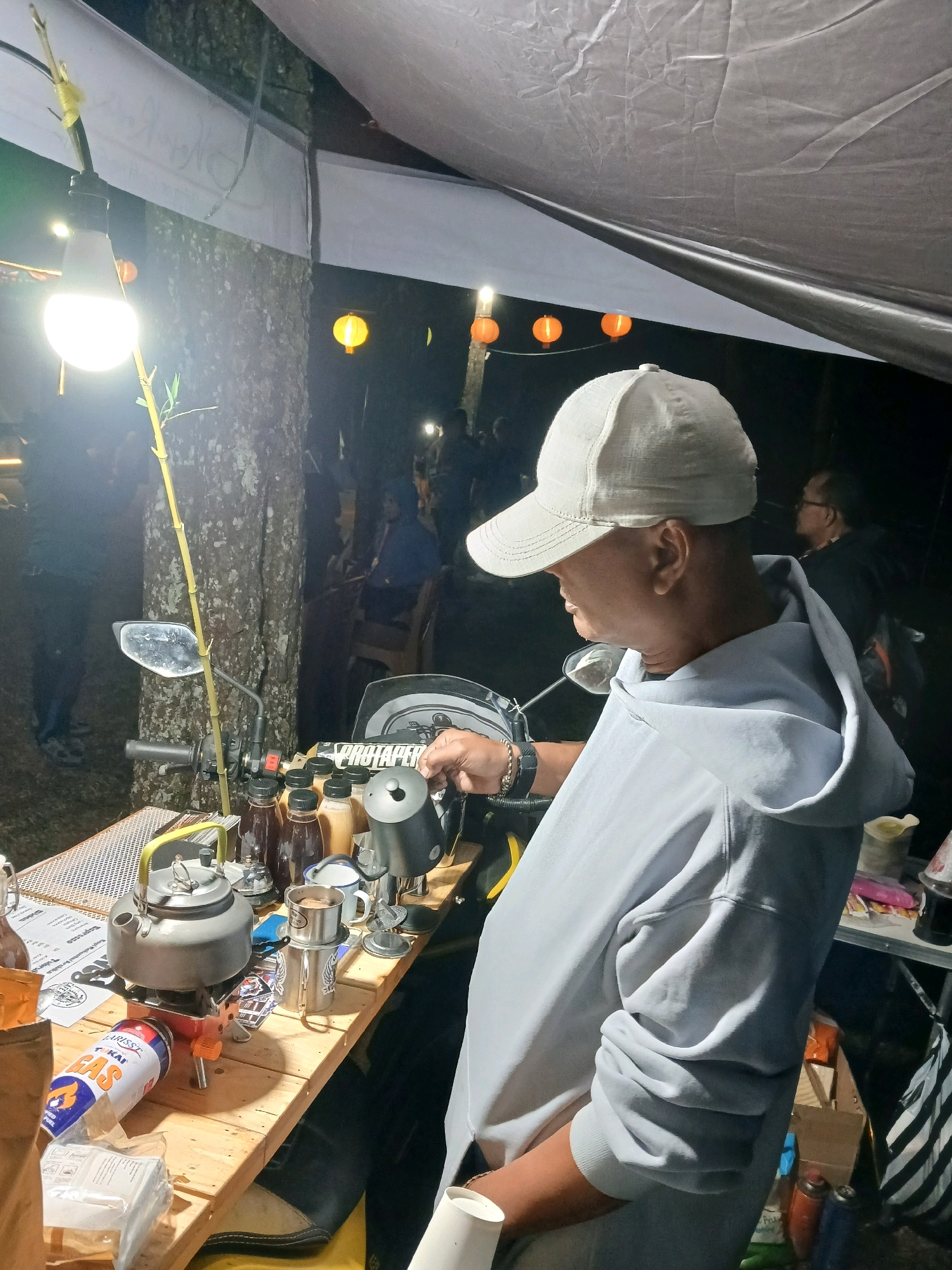 The image captures a scene at night in Berjo, Indonesia, likely a rural or natural setting given the presence of trees and an outdoor setup. A man wearing a light-colored cap and a grey hooded sweatshirt is standing behind a wooden table. He appears to be preparing a drink, possibly coffee, as indicated by the presence of a kettle, a portable stove with a gas canister, several bottles of liquid, and a metal coffee drip filter with a cup. The table is also adorned with a motorcycle mirror, suggesting a makeshift setup, possibly a mobile coffee stand.
The background is dark, illuminated by various lights, including a bright LED lamp hanging from a bamboo stalk, and several orange lanterns suspended in the distance. Other people are visible in the background, appearing as silhouettes or figures in low light, suggesting this is a public gathering or a marketplace. The overall impression is one of an outdoor nocturnal event or a coffee business operating in a relaxed, possibly camping-like, environment. The man's focus is on the task at hand, pouring liquid from a small black kettle into a coffee filter device.