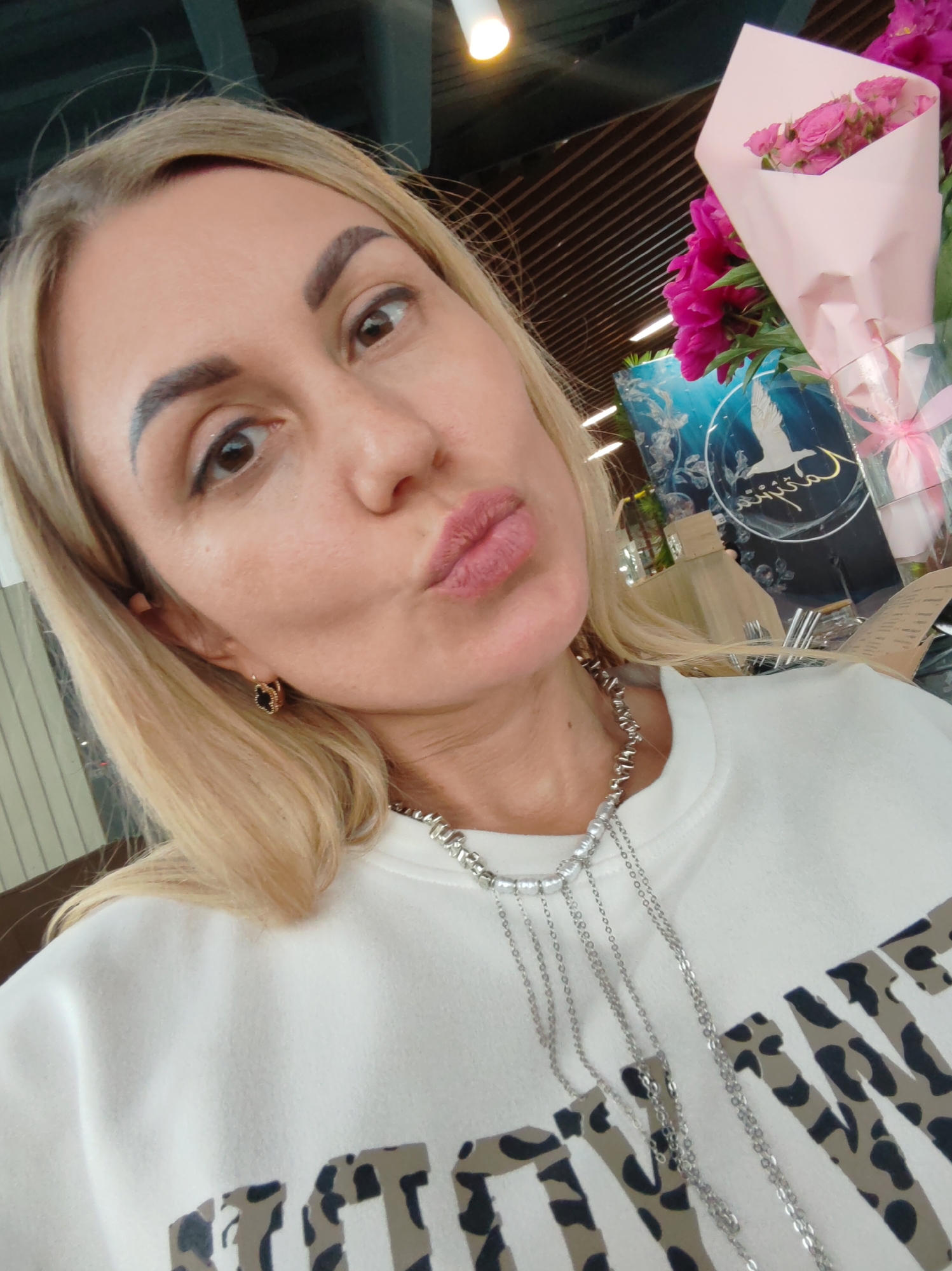 A blonde woman, with pink lipstick and large hoop earrings, looks directly at the camera with pursed lips. She is wearing a white crewneck sweatshirt with a leopard print design on the front. A silver necklace with multiple dangling chains is worn over the sweatshirt. Behind her, a bouquet of pink roses and vibrant pink peonies is partially visible, wrapped in pale pink paper. In the background, a blue circular sign with a white bird motif and text is discernable. The environment appears to be indoors, possibly a restaurant or cafe, with wooden ceiling beams and lighting fixtures visible. No specific location in Amursk, Russia can be determined from the image.