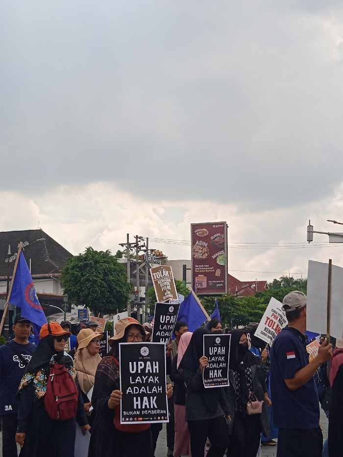 A group of people, mostly women, are marching down a street in Yogyakarta, Indonesia, which is located at -7.783 latitude and 110.367 longitude. They are carrying signs that read "Upah Layak Adalah Hak," which translates to "A living wage is a right," and "Bukan Pemberian," meaning "It's not a gift." There is a billboard in the background advertising a restaurant called "Go Ahead" featuring pictures of food, and other signs that read "Tolak Kenaikan Harga Polusi" and "Tolak Kenaikan Harga." The individuals are clad in everyday attire, including t-shirts, long-sleeved shirts, and hats. Some are carrying bags. The marchers are walking on a paved street with sidewalks. There are buildings in the background. The sky is cloudy, and the weather appears to be overcast.