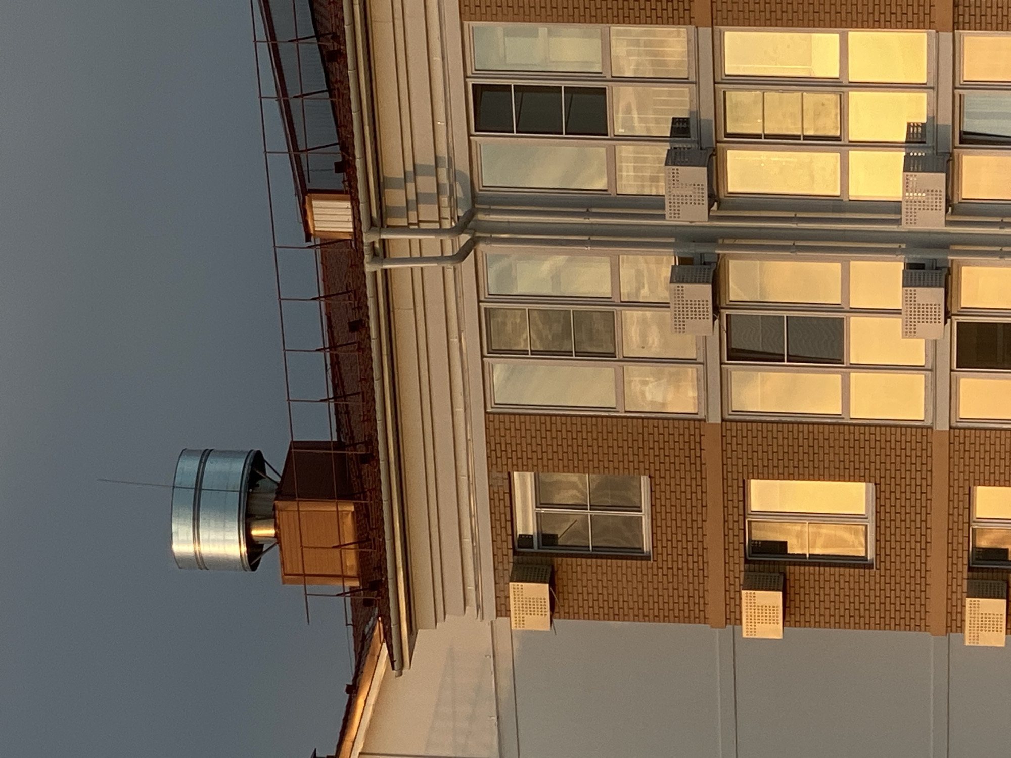 The image shows the upper floors and roof of a multi-story building, likely an apartment complex, in Dinskaya, Russia. The building has a brick facade and large windows that reflect the warm, golden light of sunset. Several air conditioning units are mounted on the exterior walls below the windows. On the roof, there is a large cylindrical vent or chimney with a metallic finish, situated on a brown wooden platform. A metal railing runs along the edge of the roof. The sky behind the building is a dark, overcast gray. There are no people visible in the image.