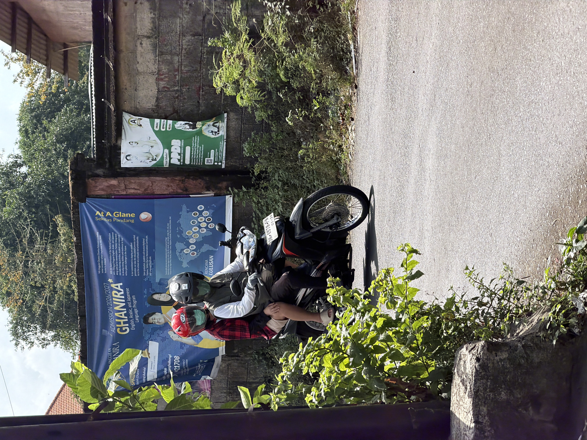 In Kuta Selatan, Indonesia, two individuals wearing helmets ride a motorcycle on an asphalt road. The motorcycle has a license plate that reads "DW-5578-FN". The rider in front wears a black helmet and a grey vest over a white long-sleeved shirt. The passenger behind wears a red helmet and a red and black checkered shirt. Both are wearing face masks. They are riding past a concrete wall adorned with several posters. One poster, a large blue banner, advertises "GHANIRA", an English, Arabic, and Japanese Language Program for admission in 2026/2027. Another poster, green and smaller, announces "PPDB", indicating a student admission process, with text in Indonesian. Lush green foliage and trees are visible in the background, with a hint of a tiled roof from a nearby building. The scene appears to be outdoors during daylight, with sunlight casting shadows on the road and surrounding vegetation.