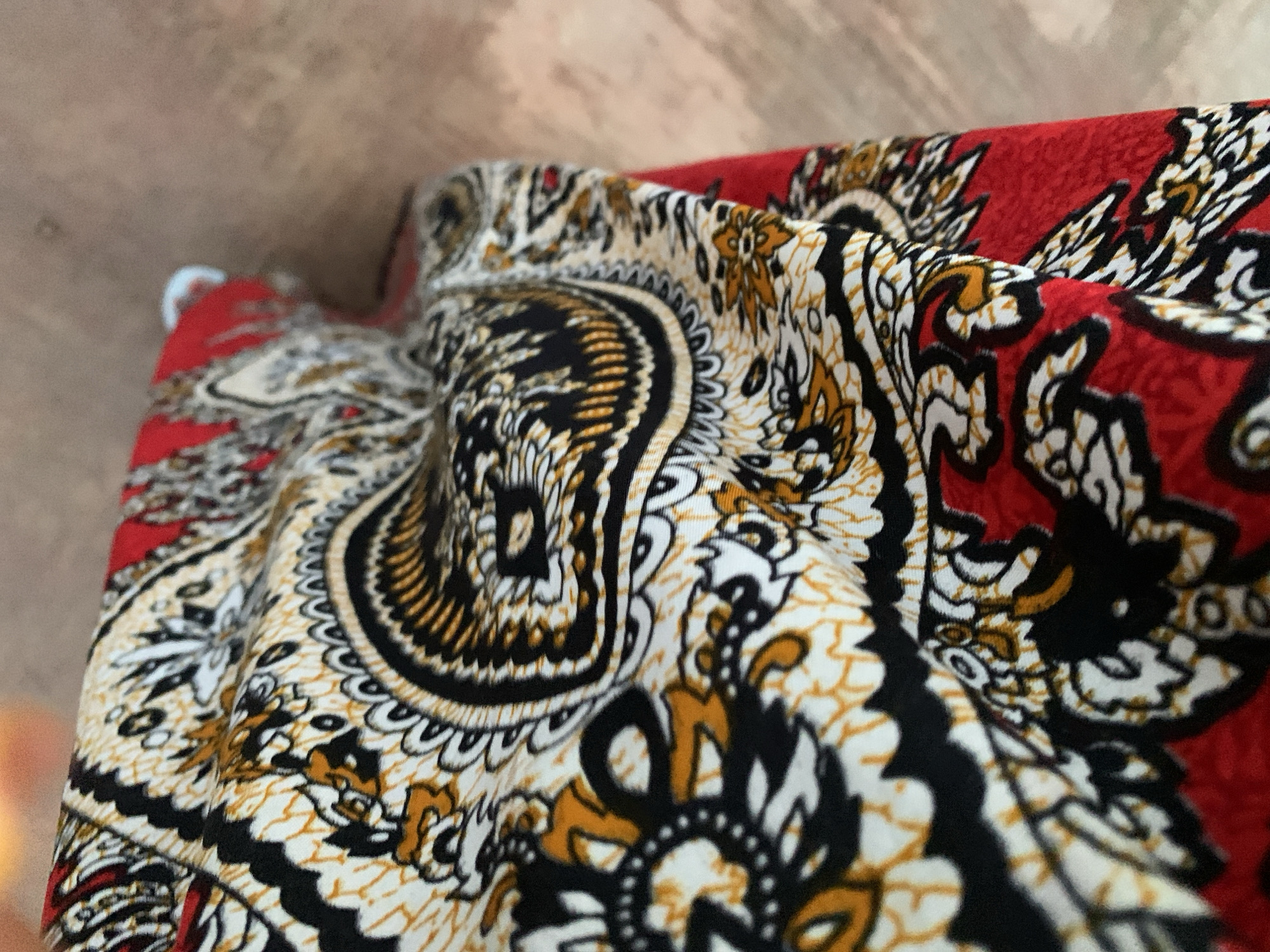 The image shows a close-up view of a piece of fabric with a paisley-like pattern. The fabric is predominantly red, white, black, and golden yellow. The pattern features intricate swirling designs with leaf-like and floral motifs. The fabric is folded, creating shadows and highlights that emphasize its texture and the dimensionality of the pattern. The background is out of focus and appears to be a light brown, textured surface, possibly a wooden table or floor. The lighting is soft and diffused, highlighting the colors and details of the fabric.
