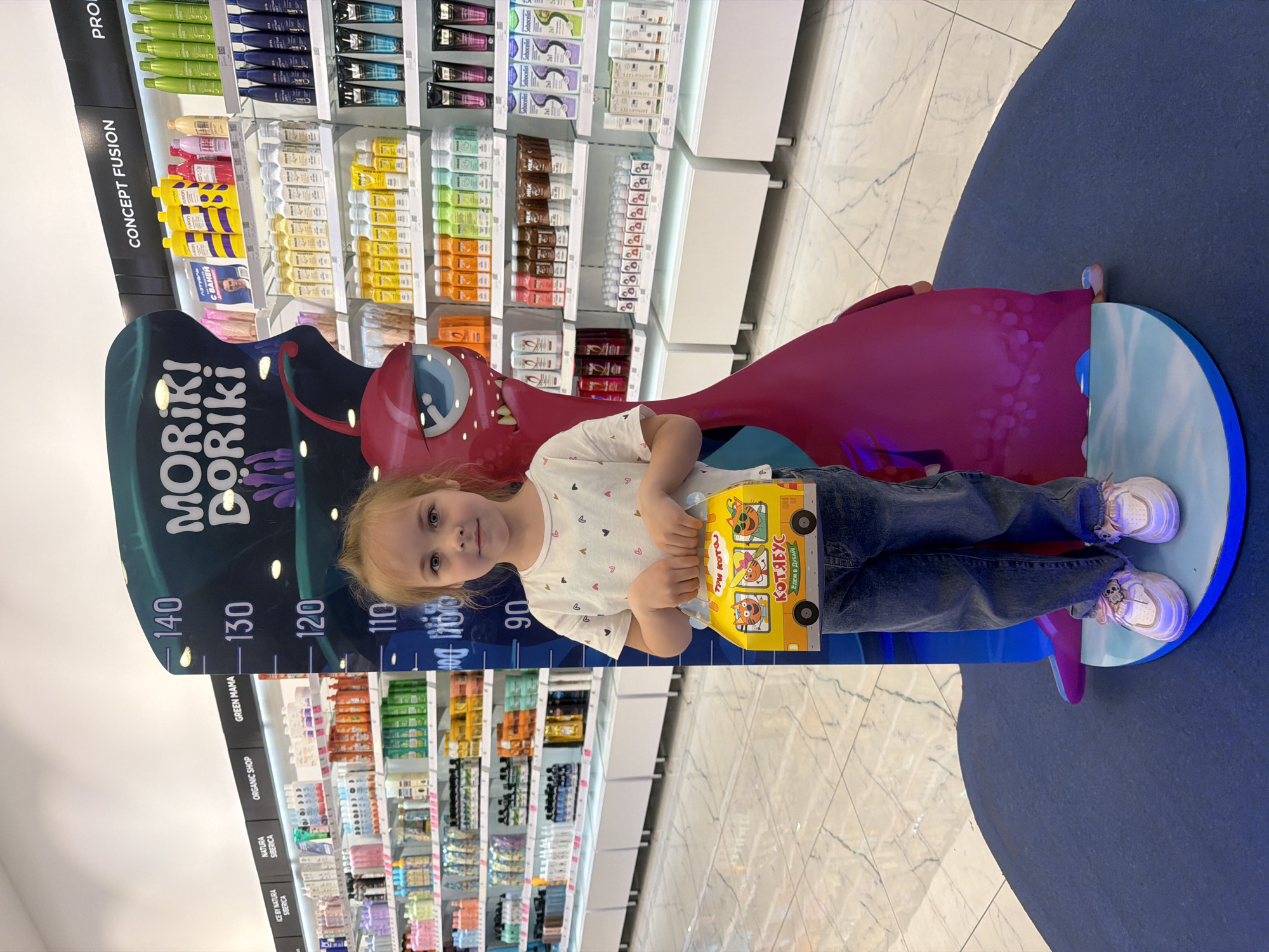 A young girl stands on a blue and white platform shaped like a circle, positioned next to a height chart with a cartoon monster figure. The chart measures height in centimeters from 90 to 140. She is wearing a white t-shirt with small heart patterns, blue jeans, and white sneakers. The girl is holding a yellow box with cartoon cats printed on it, with the text "ТРИ КОТО КОТЯБУС", which translates to "Three Cats Kotyabus", and "ЕДЕМ В ДУБАЙ", meaning "We are going to Dubai".
Behind her, shelves stocked with numerous hair products are visible. The shelves are labeled with brand names such as "CONCEPT FUSION", "PRO", "GREEN MAMA", "ORGANIC SHOP", "NATURA SIBERICA", and "ICE BY NATURA SIBERICA".
The scene is brightly lit, and the floor is patterned with light-colored tiles. The location is a retail store in Novoaleksandrovka, Russia, likely a cosmetics or personal care store.