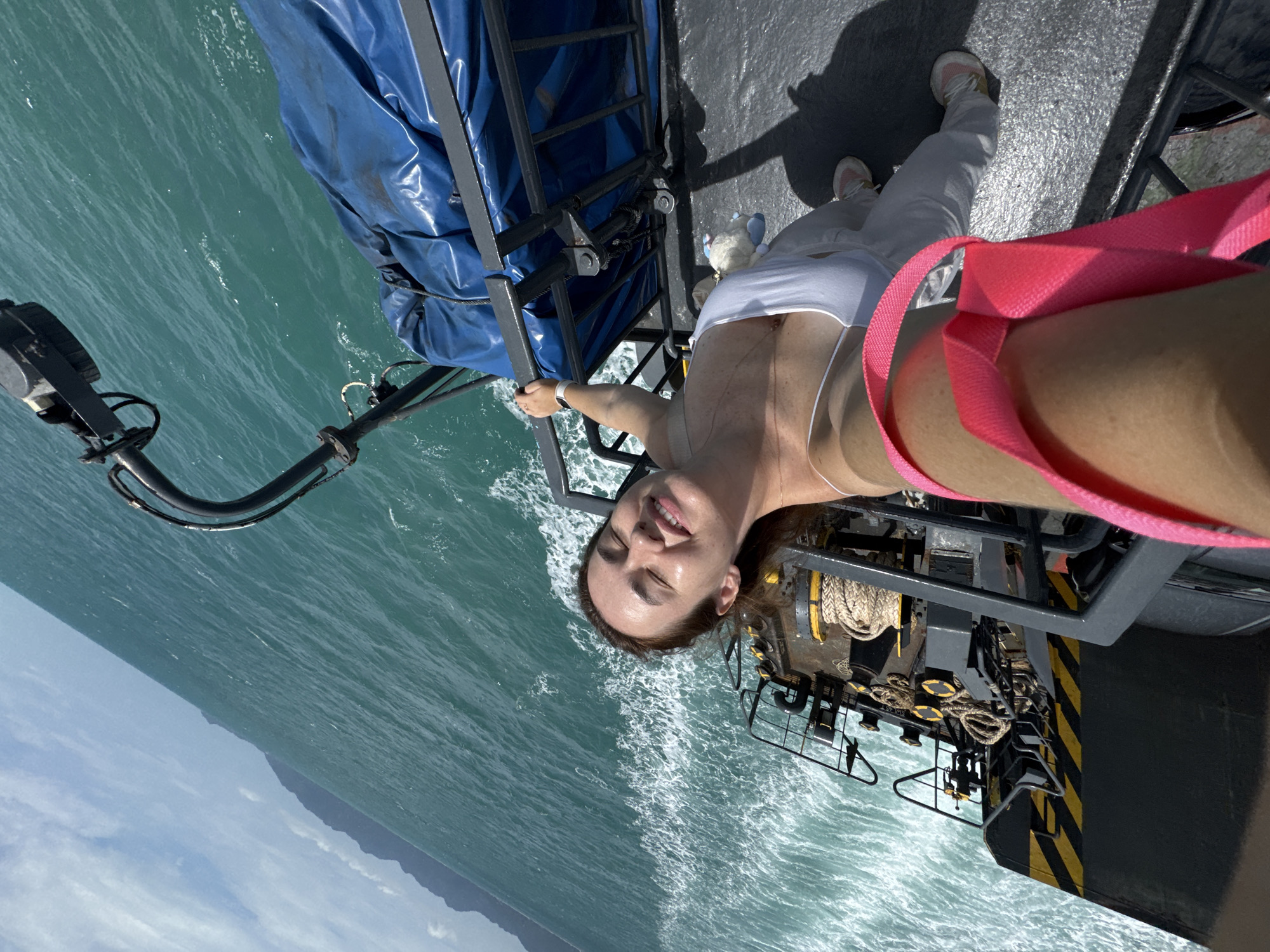 A woman is taking a selfie on the deck of a ferry in the ocean off the coast of Ko Pha-ngan, Thailand. She is smiling with her eyes closed, her head tilted back. The water is a clear turquoise, and waves are visible behind her. A blue tarp covers cargo on the ferry. The woman is wearing a white tank top and white pants. She is holding onto a metal railing with her left hand. The camera is angled downwards, capturing her face and upper body, as well as the deck of the ferry and the ocean. The ferry appears to be moving, as evidenced by the wake behind it. The sky is blue with some white clouds. In the distance, a mountainous coastline is visible.