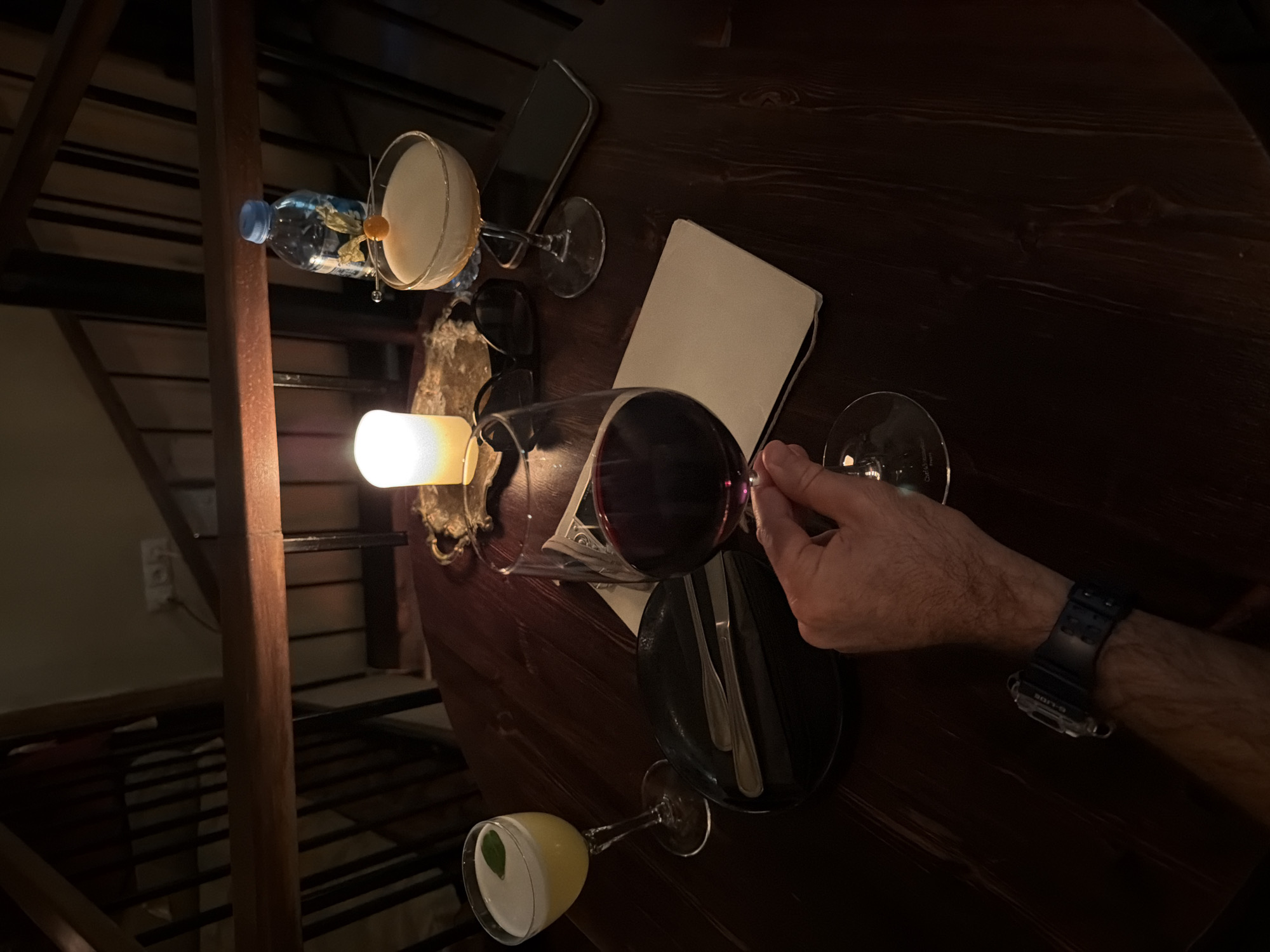 The image depicts a dimly lit scene at a table in what appears to be a restaurant or bar, potentially in Tbilisi, Georgia. The table is made of dark wood. Visible on the table are two cocktail glasses (one with a white drink and the other with a yellow drink with a basil leaf), a glass of red wine being held by a hand, a phone, sunglasses, a notepad, silverware on a black plate, and a water bottle. A lit candle is present, adding to the ambient lighting. A hand with a black wristwatch is holding the wine glass. In the background, a staircase structure is partially visible.