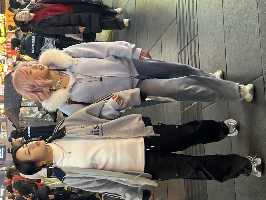Two East Asian females walk side-by-side on a paved pedestrian street in Yuzhong District, China, under artificial evening lighting. The female on the left, with dark hair, wears a white mock-neck shirt, a grey bomber jacket with navy accents and visible text "nis", black cargo pants with silver-colored buttons and drawstrings, and silver athletic shoes. She has layered silver necklaces. Her right hand is linked with the left hand of the female on her right. This second female, with short wavy pink hair, wears a white inner shirt, a light blue zipped hoodie featuring a prominent white faux fur collar and a small yellow emblem on the chest, light-wash blue jeans, and cream-colored athletic shoes. She wears a colorful beaded necklace and black wired earphones around her neck. Both females are looking generally forward and slightly rightward. The background shows a crowded urban environment with numerous blurred pedestrians, illuminated commercial signage, a dark canopy structure, and a partial view of a scooter. The ground is composed of grey square tiles with horizontal tactile strips.
