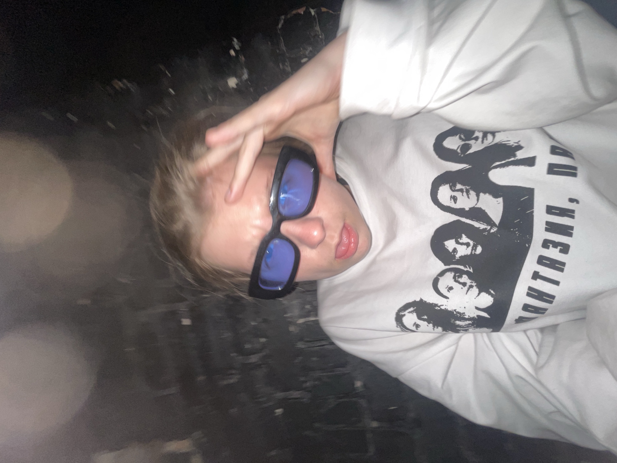 A young person is depicted lying down, angled upwards and to the right, in a dimly lit or dark environment. The subject is fair-haired, seen from the upper torso, wearing black rectangular sunglasses with blue reflective lenses, and a long-sleeved white t-shirt. One hand is raised to their forehead, partially obscuring the top of their head. Their lips are slightly parted, revealing a glimpse of their tongue.
The activity appears to be a casual pose, possibly a selfie taken in a relaxed or playful manner. The background is mostly dark and indistinct, suggesting an indoor setting with low light or a dark outdoor area at night. There are some faint, blurry light flares in the foreground and background, indicative of light sources or reflections in the dark.
Notable details include the subject's expression, which is obscured by the sunglasses but suggests a relaxed or slightly playful demeanor. The overall lighting is low-key. The location cues are limited to the text on the t-shirt.
The white t-shirt features a black graphic print of several stylized faces, resembling a band or group. To the right of these faces, partial Cyrillic text is visible. The legible parts read "ФАНТАЗИЯ, ПР...". This transliterates to "FANTAZIYA, PR...", likely referring to the Russian band "ФАНТАЗИЯ ПРЕДЕЛА" (Fantaziya Predela), which means "Fantasy of the Limit." Given the context of Moscow, Russia, this is a plausible cultural reference.