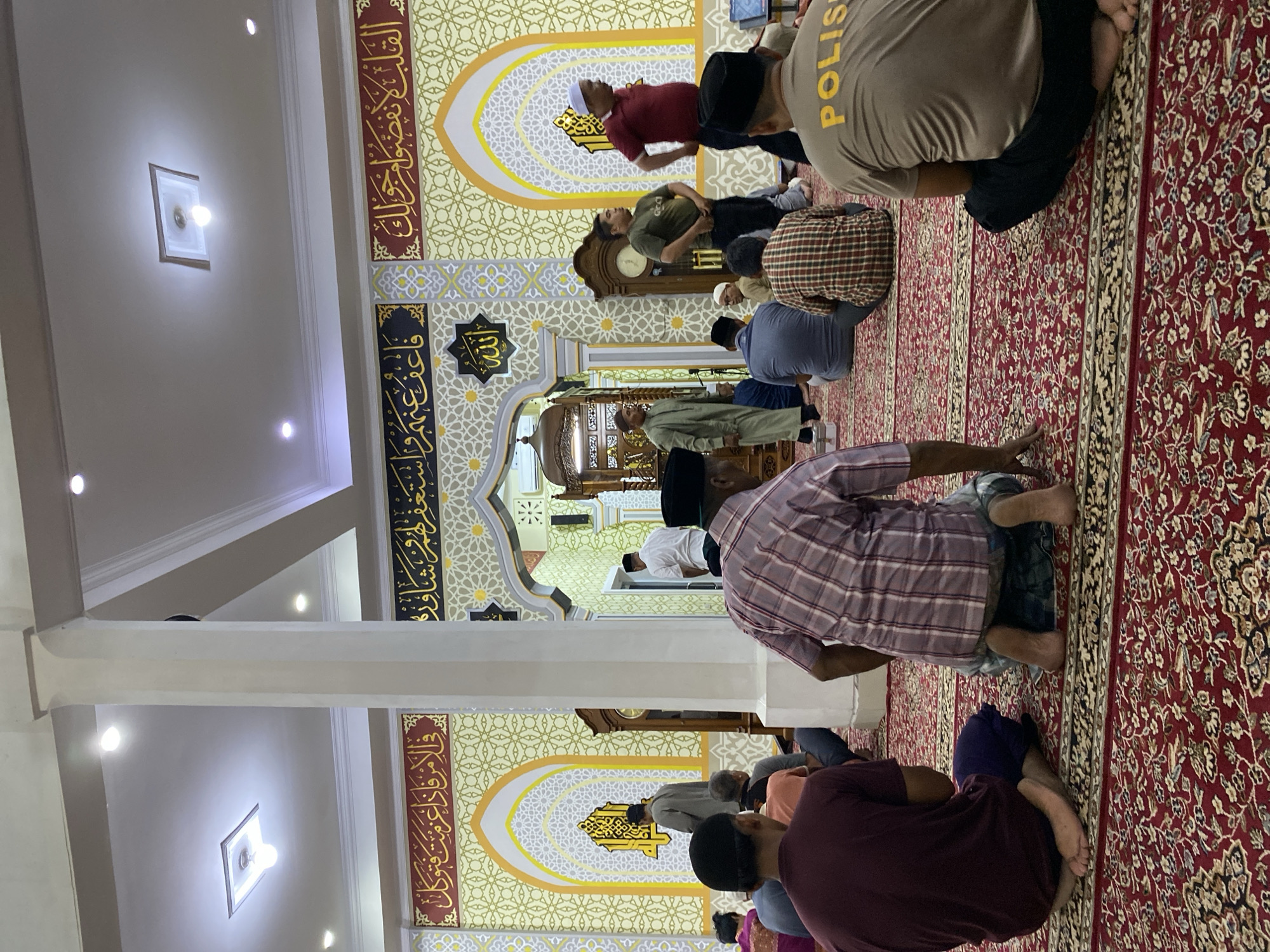 The image depicts the interior of a mosque in Lhokseumawe, Indonesia, with several men engaged in prayer or observing. The setting is ornately decorated with Islamic calligraphy and geometric patterns on the walls. The flooring is covered with a large, intricate red carpet. In the foreground, the backs of several men are visible, some in prayer positions. One man in the middle ground is wearing a shirt with the word "POLIS" emblazoned on it in yellow. Other individuals are seen standing or sitting, dressed in traditional Islamic attire and casual clothing. The mosque features tall white pillars and recessed lighting in the ceiling. A wooden grandfather clock is positioned against one of the walls. The overall atmosphere suggests a communal religious gathering.