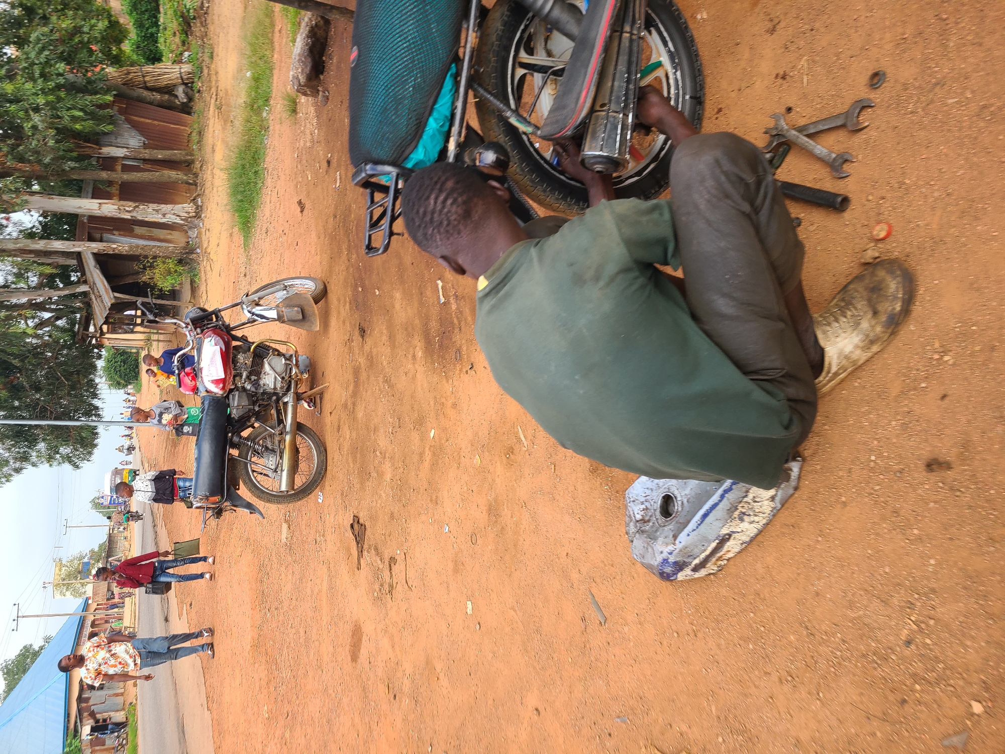 In Vom, Nigeria, a man is crouched on reddish-brown dirt, working on the rear wheel of a motorcycle. He wears a dark green shirt and dark pants. Tools, including wrenches, lie on the ground near him. A detached motorcycle fuel tank, a blue and silver metal object, rests on the dirt to his left. Behind him, another motorcycle, predominantly red with black accents, is parked on the dirt. In the background, a street runs horizontally, with people walking on the left side. A building with a blue roof and other structures line the street, amidst trees and vegetation. The sky is overcast.