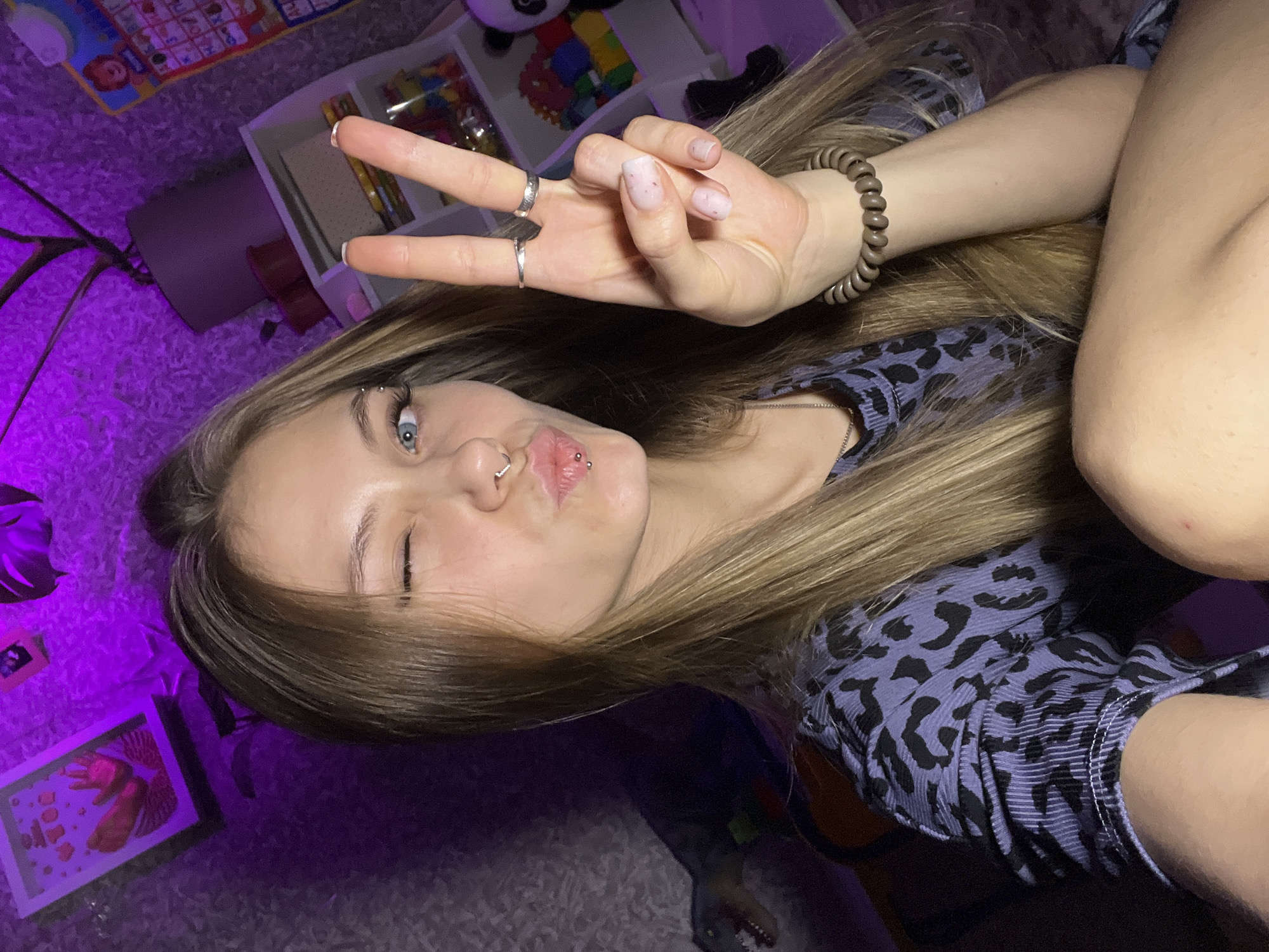 A young woman is lying on a carpeted surface, partially lit by a purple light source. She is wearing a shirt with a blue and black animal print and has blonde hair, light blue eyes, and multiple piercings in her face, including her eyebrow and lip. She is making a peace sign with her hand, which has a hair tie bracelet on the wrist and rings on two fingers. In the background, there are toy storage containers, a panda plush, and an alphabet poster. The location of Фоки, Russia is not discernible from the image.