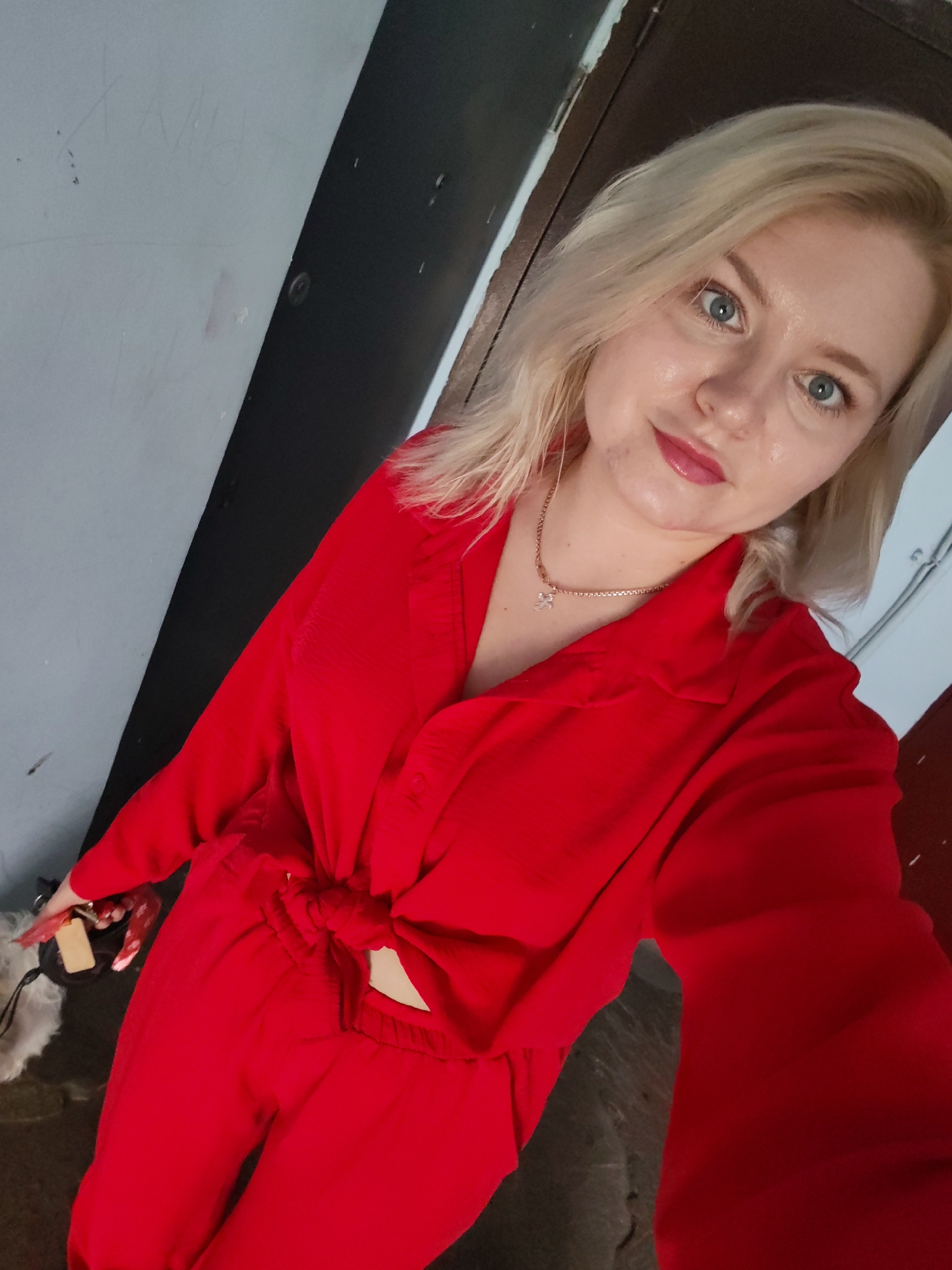 A woman with blonde hair and blue eyes is taking a selfie in a dimly lit hallway with a gray wall on the left and a dark door on the right. She is wearing a red collared shirt tied at the waist and matching red pants. She is also wearing a delicate gold necklace with a small pendant. Her left hand is visible holding a black bag with a fluffy white trim and a small tag. The wall on the left has faint, scrawled markings. The scene appears to be an indoor residential or public hallway. There is no specific information to place this image in Izhevsk, Russia.