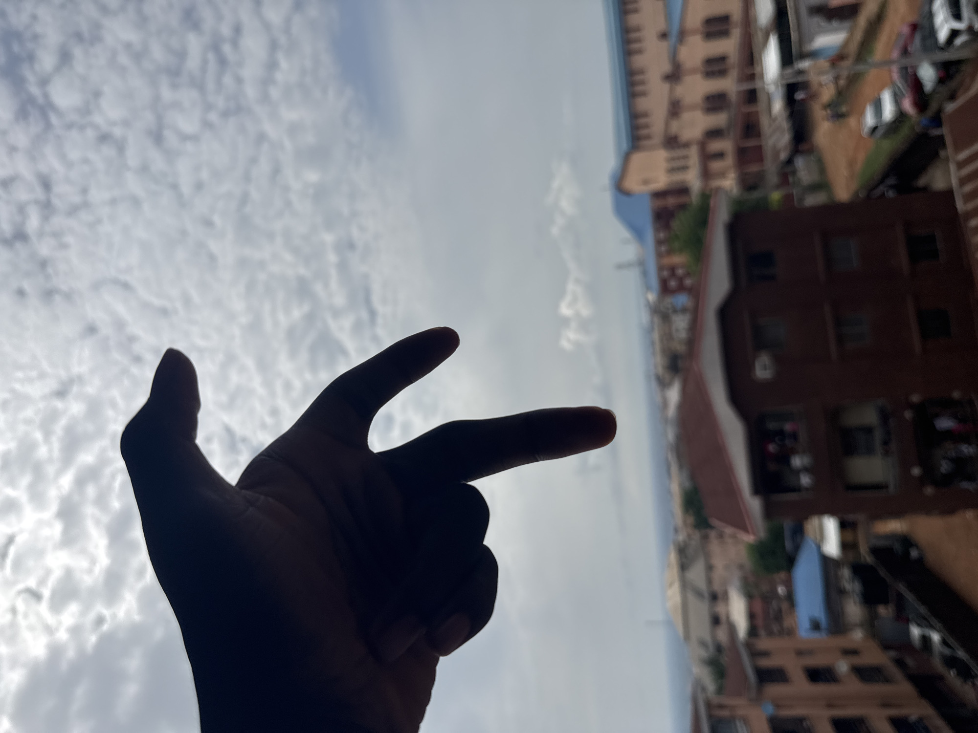 The image shows a hand silhouetted against a background of clouds and urban buildings. The hand is making a gesture resembling the shape of a gun with the index and middle fingers extended. The buildings, of varying heights and colors primarily shades of brown and beige, suggest an urban setting. Cars are also visible, indicating streets. The scene appears to be captured from an elevated position, possibly from a high floor. The geographical location is specified as Onitsha, Nigeria.