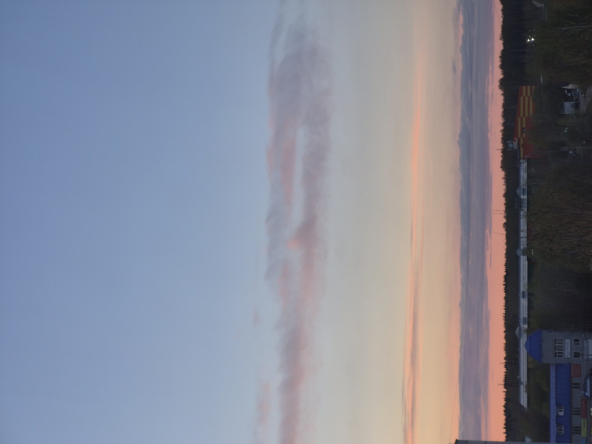 The image shows a sunset in Tobolsk, Russia. A tall blue and gray building is visible in the foreground. Behind the building is a line of trees. Beyond the trees, the sky transitions from blue to pink and orange hues with striated cloud formations. A red and yellow building stands out along the tree line. Power lines can be seen above the trees.