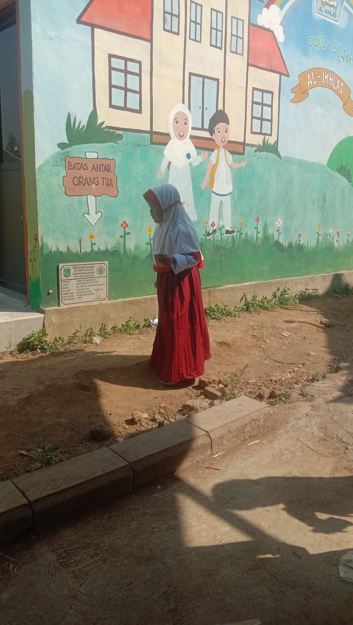A young girl wearing a hijab and a long red skirt stands in front of a colorful mural. The mural depicts a school building with cartoon children and a banner that reads "PAUD - ISLAM AL - IKHLAS". Another banner on the mural reads "BATAS ANTAR ORANG TUA", meaning "Parent Drop-off Limit". A small plaque with text in Indonesian is affixed to the wall below the mural. The girl is looking down at a book or papers in her hands. The ground is dirt and concrete, with some scattered debris. The scene is outdoors, with shadows cast by the sun, suggesting it is daytime. The environment appears to be a schoolyard in Karanganyar, Indonesia, based on the text in the mural and the plaque.