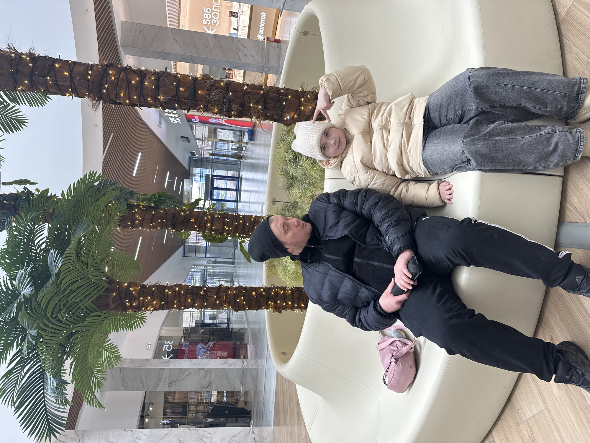 The image shows an indoor scene, likely in a shopping mall in Izhevsk, Russia. A man and a girl are resting on a curved white bench. The girl is lying down, wearing a beige puffer jacket, a white hat, and grey jeans. She is gesturing with her hand near her head. The man is seated next to her, dressed in a black puffer jacket, a black hat, and black athletic pants. He is holding a phone or similar device. A pink backpack sits next to him on the bench.
Artificial palm trees with decorative lights line the corridor behind the bench. Shopfronts are visible in the background. Signs written in Russian are also visible, suggesting the location. The flooring is a mix of light wood and marble patterns.