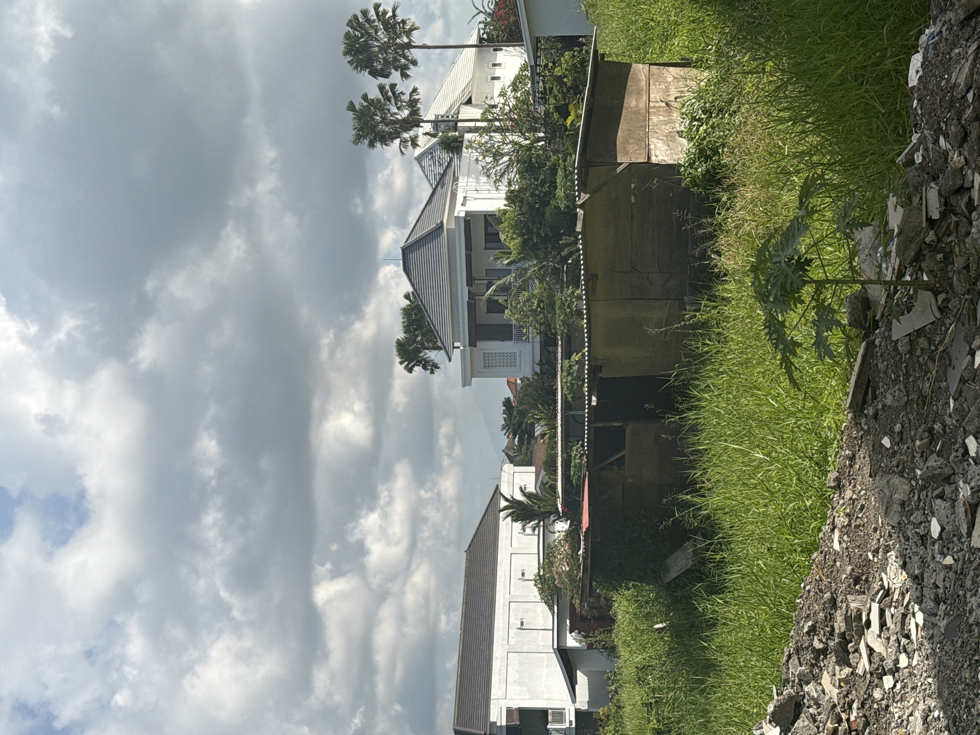 The image shows a residential area in North Kuta, Indonesia under a cloudy sky. Several white multi-story buildings with gray roofs are visible, interspersed with palm trees and lush greenery. A makeshift fence runs along the edge of the properties. In the foreground, there is overgrown grass and a pile of rubble consisting of broken concrete and other debris. No people are visible.