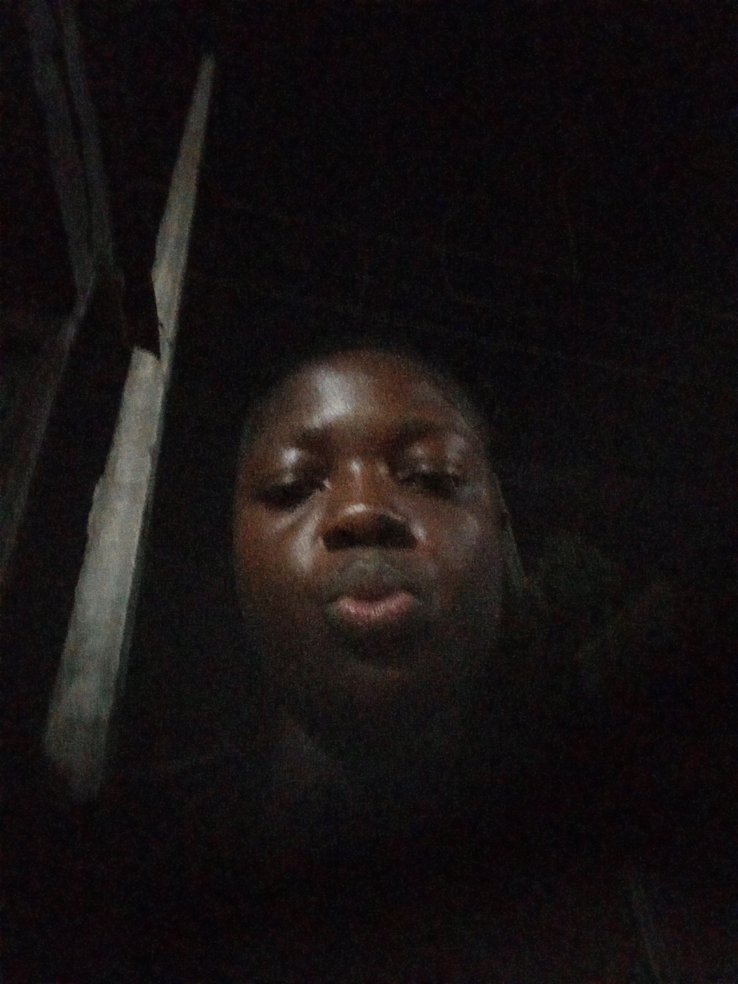 A close-up selfie of a person with dark skin is the main subject of the image. The person is looking directly at the camera with a neutral expression. The background is very dark, making it difficult to discern any specific details. A blurry object resembling a pole or piece of wood is visible on the left side of the frame. It is difficult to ascertain further details or location in Mbribit, Nigeria due to the poor lighting and limited visibility.