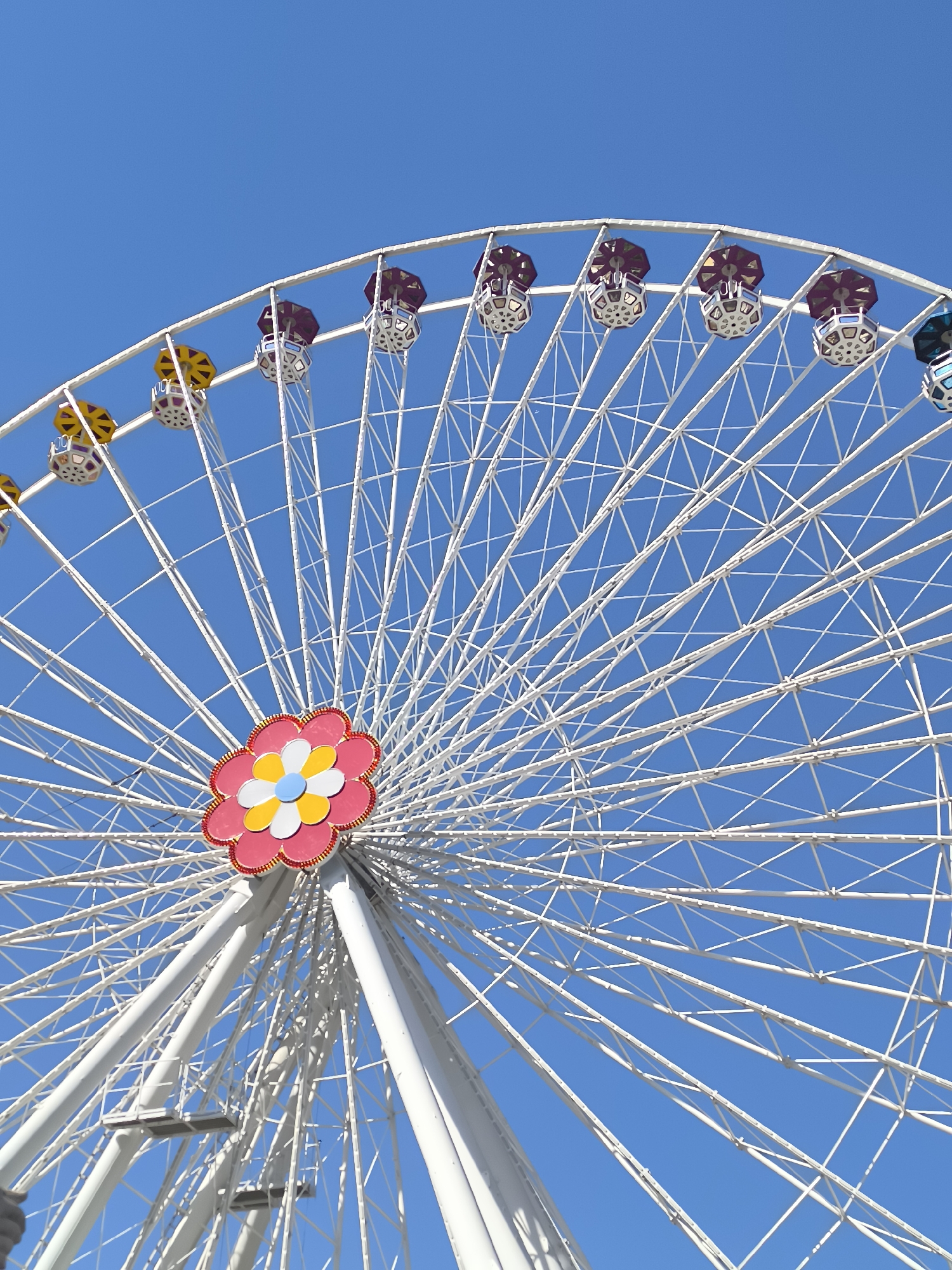 A close-up, low-angle shot shows the upper portion of a white Ferris wheel against a bright blue sky. The Ferris wheel features numerous spokes radiating from the center, forming a complex geometric pattern. Several gondolas, some with yellow umbrellas and others with purple tops, are visible attached to the rim. In the center of the wheel is a colorful, stylized flower design with pink petals, yellow and white inner petals, and a light blue center. The image appears to be taken in Vienna, Austria, likely at the Prater amusement park, known for its iconic Giant Ferris Wheel. There are no people visible in the frame, and the scene is static.