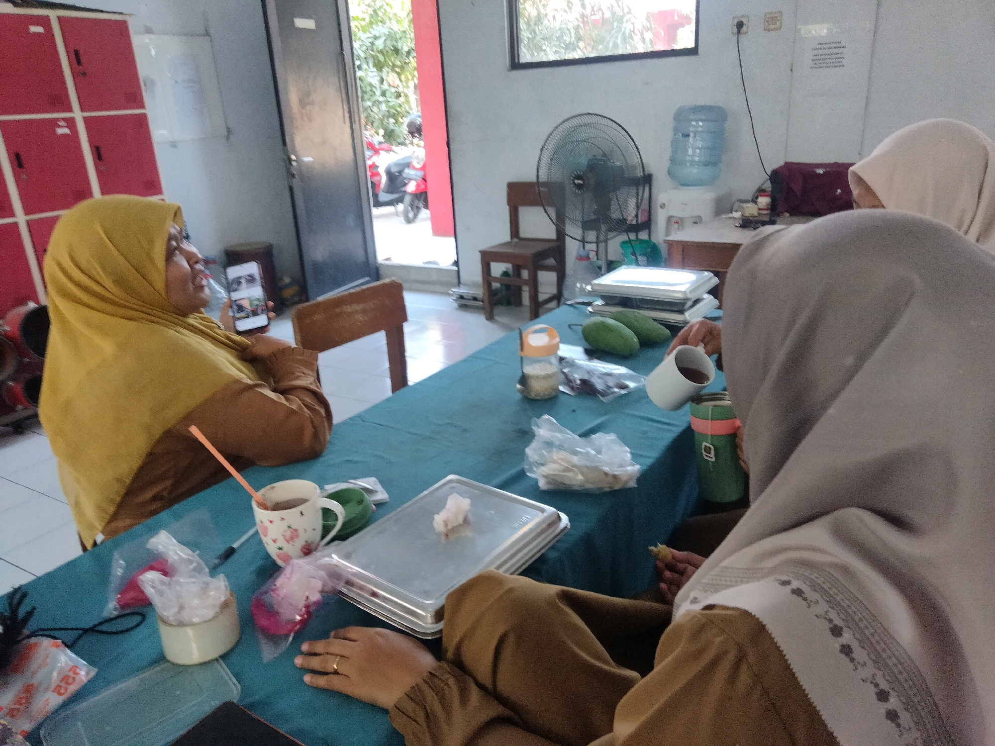 In Suka Sari, Indonesia, an indoor scene depicts three individuals, two of whom are wearing hijabs, gathered around a blue-clothed table. One woman in a yellow hijab is holding a smartphone, seemingly looking at it. Another person, mostly obscured by a light gray hijab and brown garment, is seated and appears to be holding a white mug. A third individual, also wearing a hijab, is partially visible in the background. The table is laden with various items, including a white mug with a floral pattern, a green thermos, plastic bags containing unknown contents, a metal tray with a piece of food, and other miscellaneous objects. In the background, a black oscillating fan stands next to a wooden chair. A water dispenser is also visible, along with red lockers and an open doorway leading to an outdoor area where a red motorcycle is parked. The environment appears to be a casual gathering space.